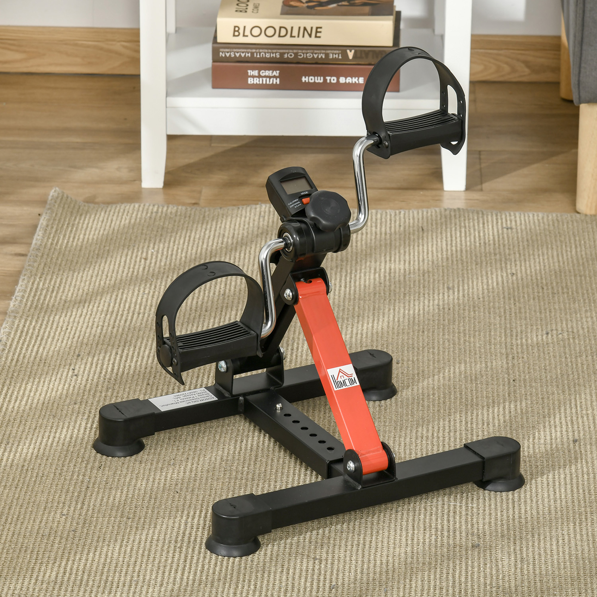 Una bicicleta de ejercicio sobre una alfombra beige. Tiene pedales negros y un soporte rojo. El fondo incluye una estantería y libros.
