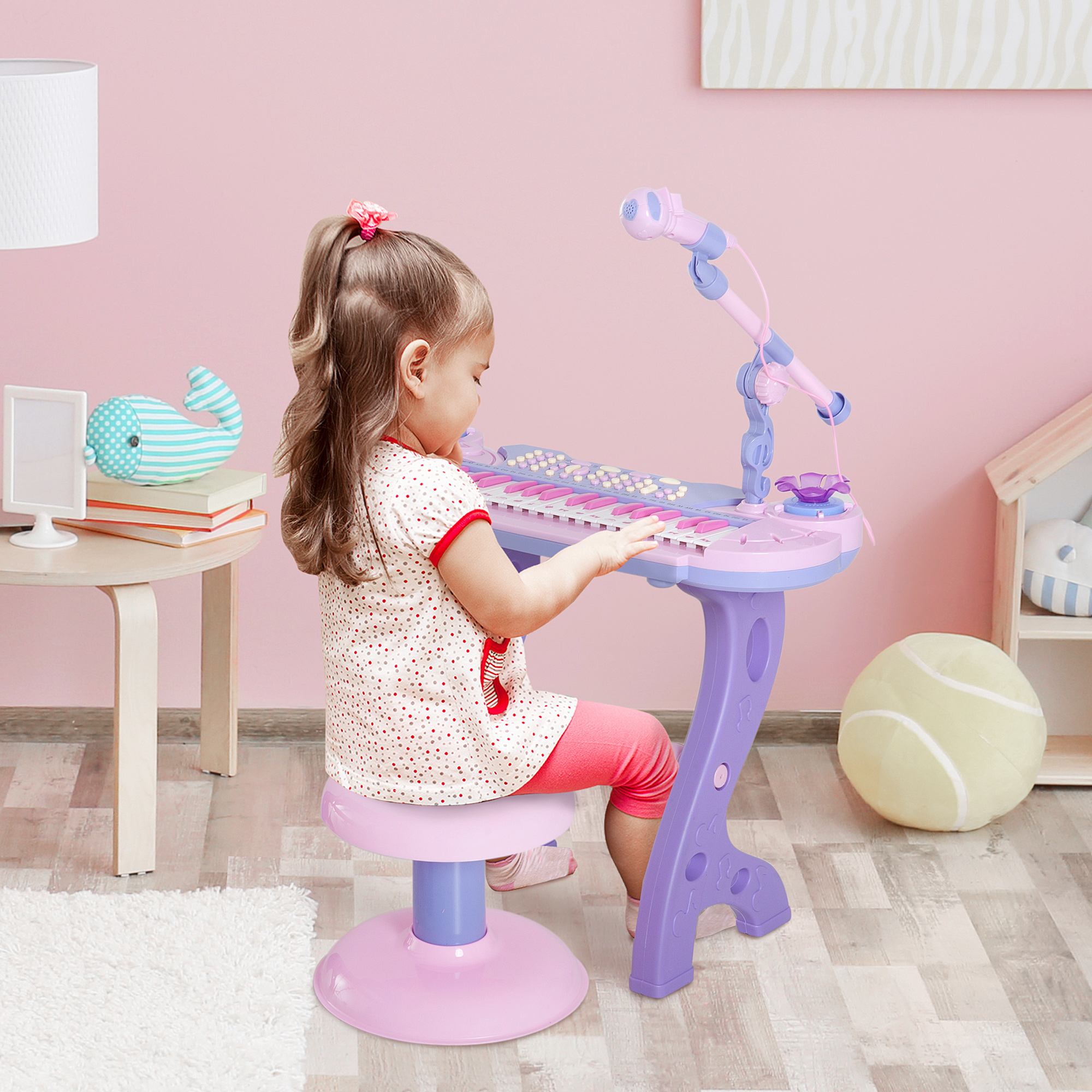 Una niña toca un piano de juguete. Lleva una camisa de lunares y pantalones rosas. Un micrófono está frente al piano.