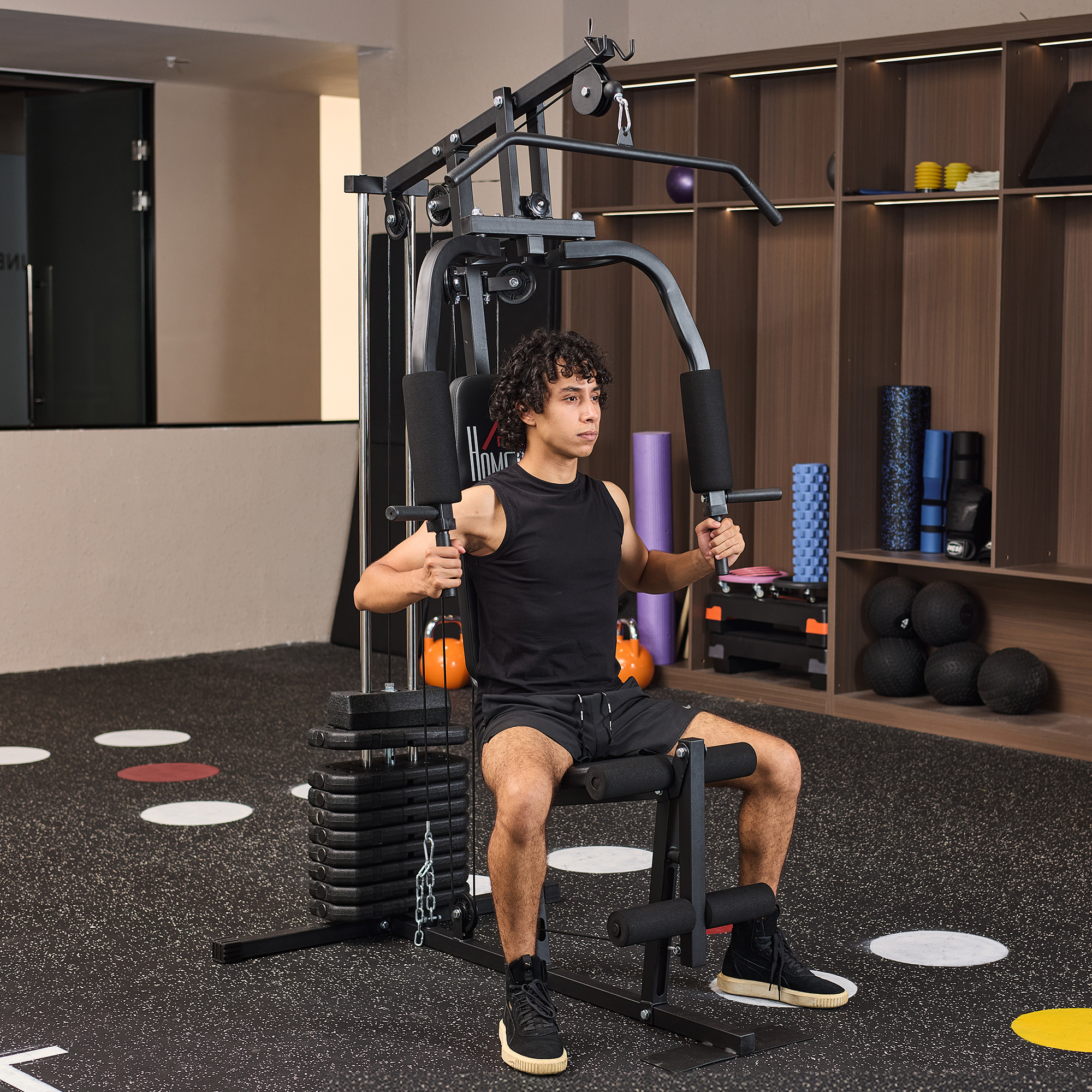 Un hombre está haciendo ejercicio en una máquina de gimnasio. Lleva ropa deportiva negra. El fondo tiene equipo de gimnasio.
