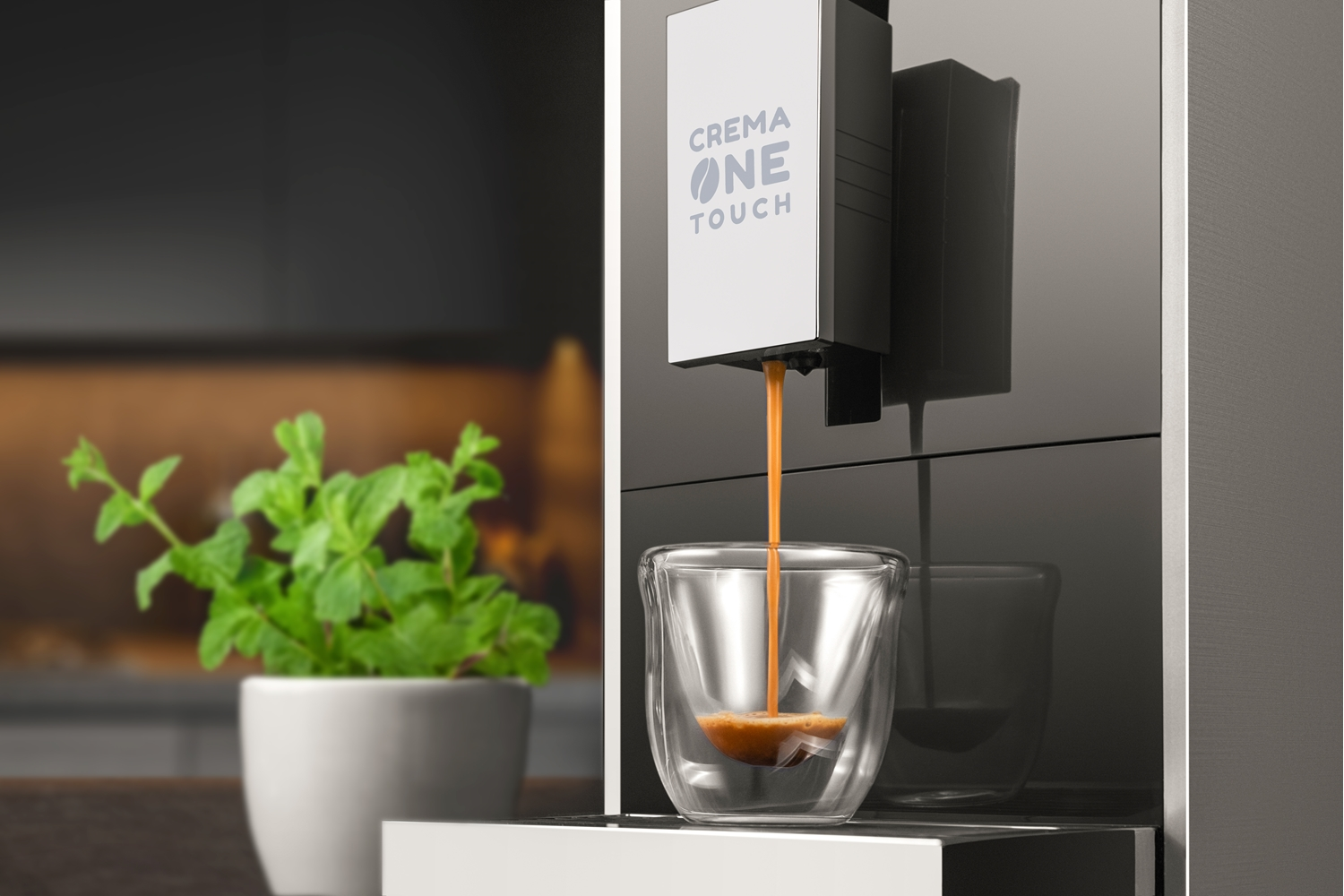 Macchina da caffè che eroga espresso in un bicchiere di vetro. Una pianta in vaso a sinistra. La macchina ha la scritta "CREMA ONE TOUCH".