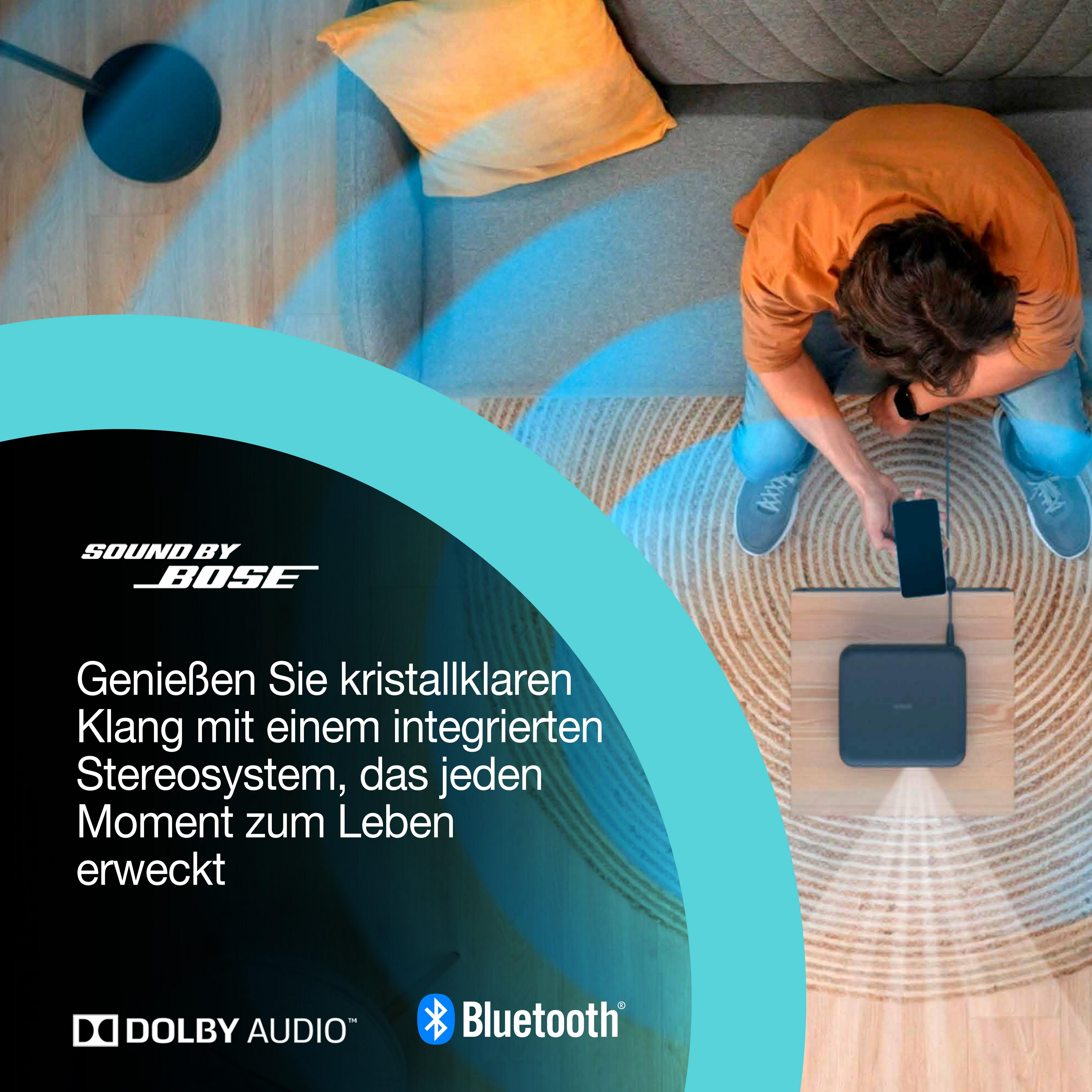 Osoba używa smartfona do sterowania czarnym urządzeniem. Osoba siedzi blisko sofy, nad urządzeniem.