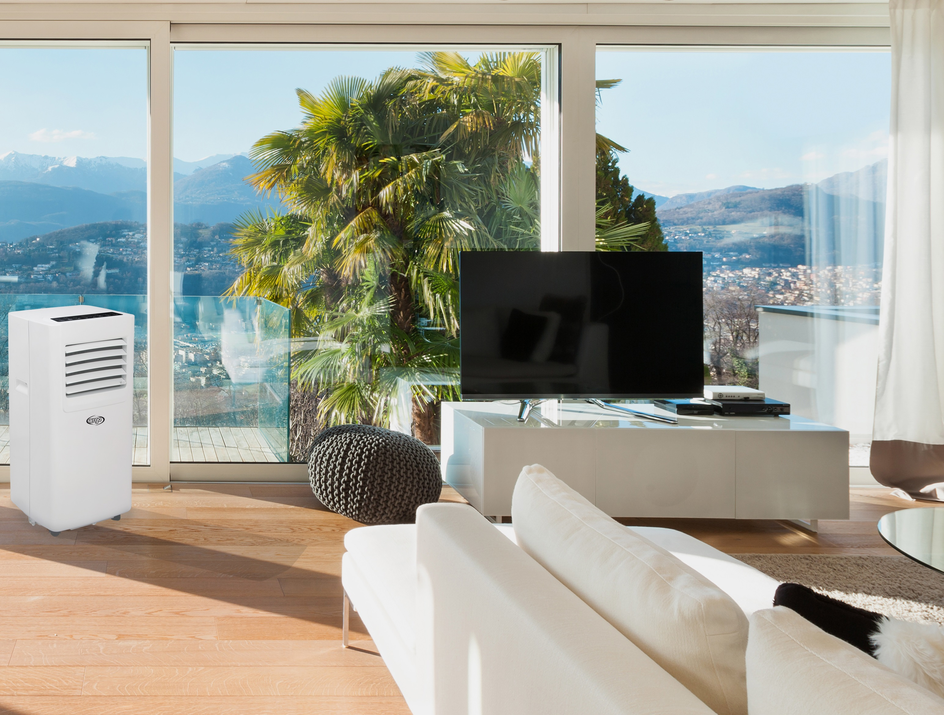 Soggiorno con condizionatore bianco, TV, divano bianco e ampie finestre con vista sulle montagne e palme.