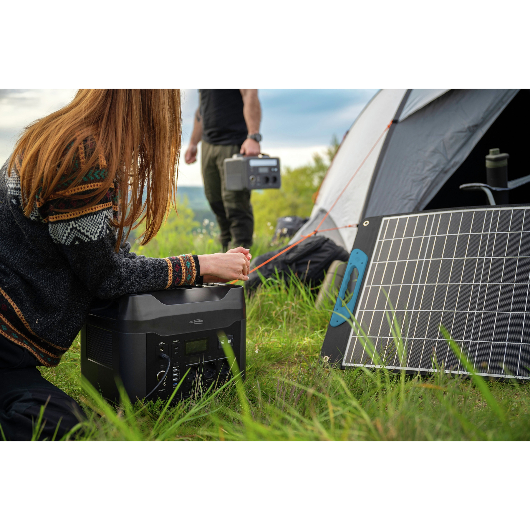 Kobieta klęczy obok elektrowni. Mężczyzna trzyma mniejszą elektrownię. Namiot i panel słoneczny w tle.