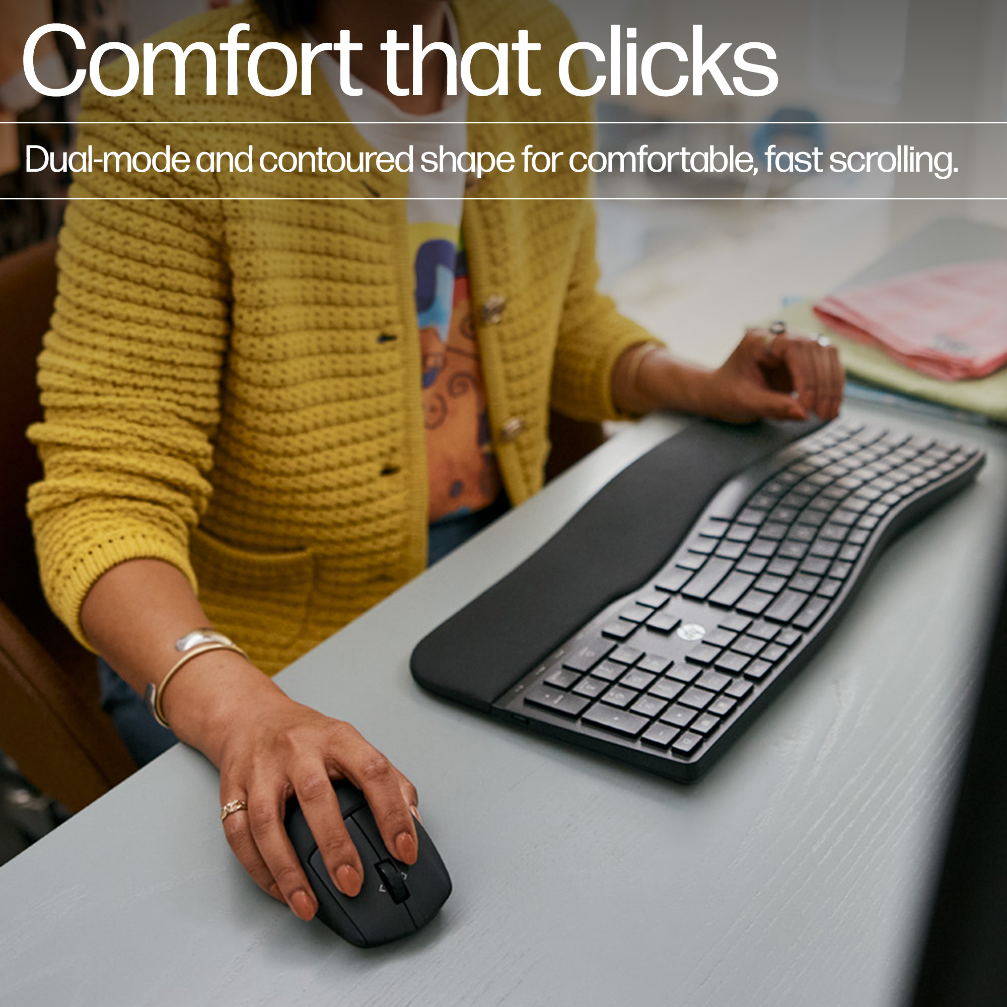 Una persona usa un ratón negro y un teclado ergonómico. La persona lleva un jersey amarillo y anillos. El teclado es curvo.