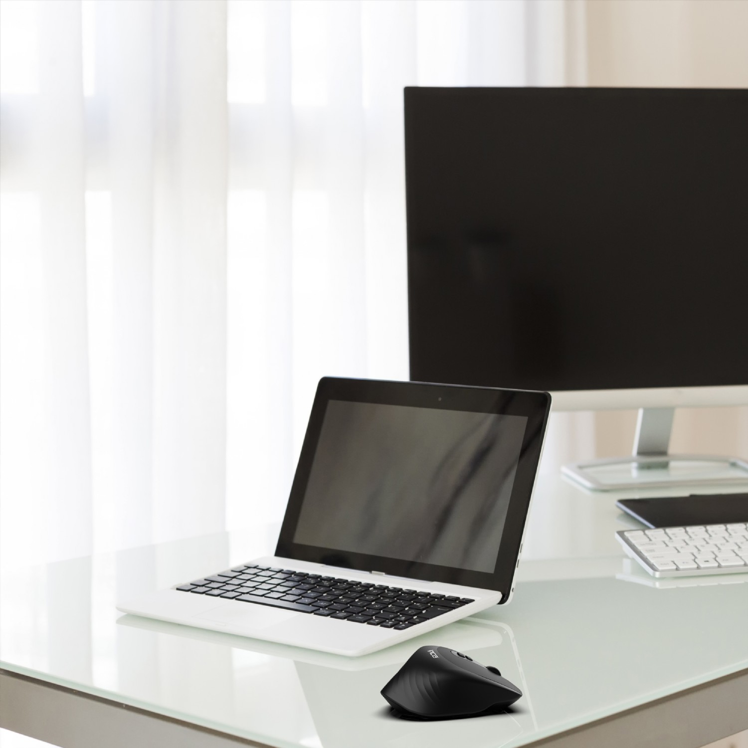 A laptop and a computer mouse sit on a table. A computer monitor and a keyboard are in the background.