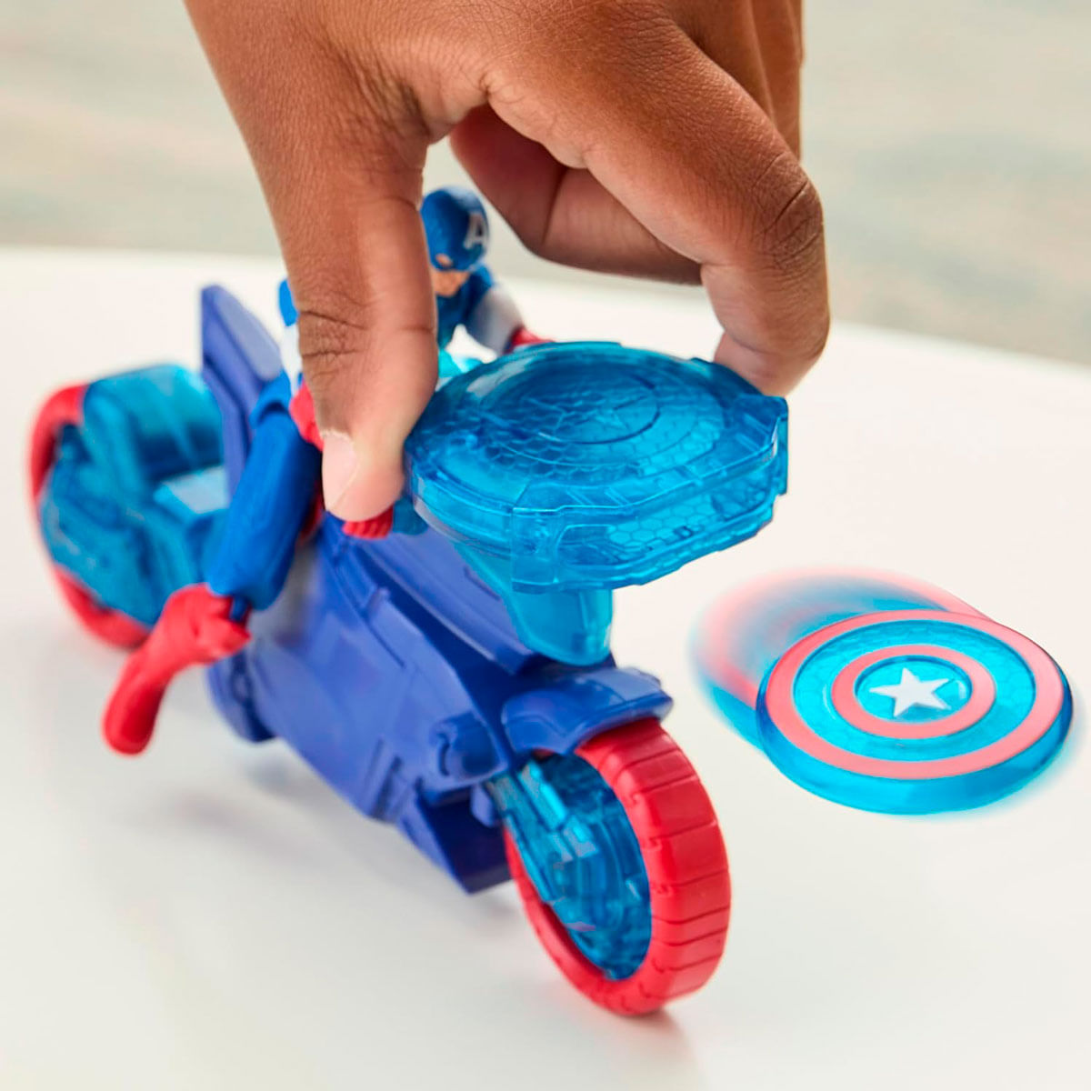 Ręka trzymająca zabawkę Kapitana Ameryki na niebieskim motocyklu. W pobliżu tarcza. Motocykl ma czerwone koła, tarcza gwiazdę.