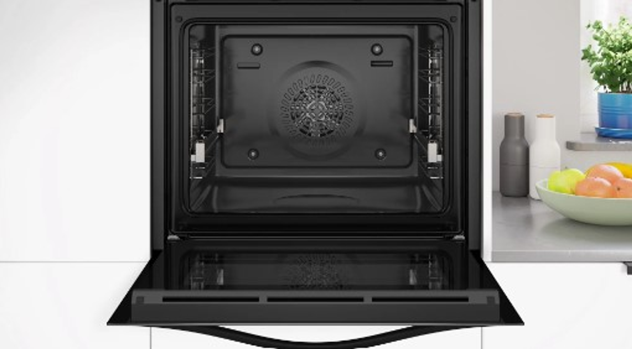 Otwarty piekarnik, czarne wnętrze, otwarte drzwi. Owoce i butelki na blacie po prawej. Białe i nierdzewne powierzchnie.