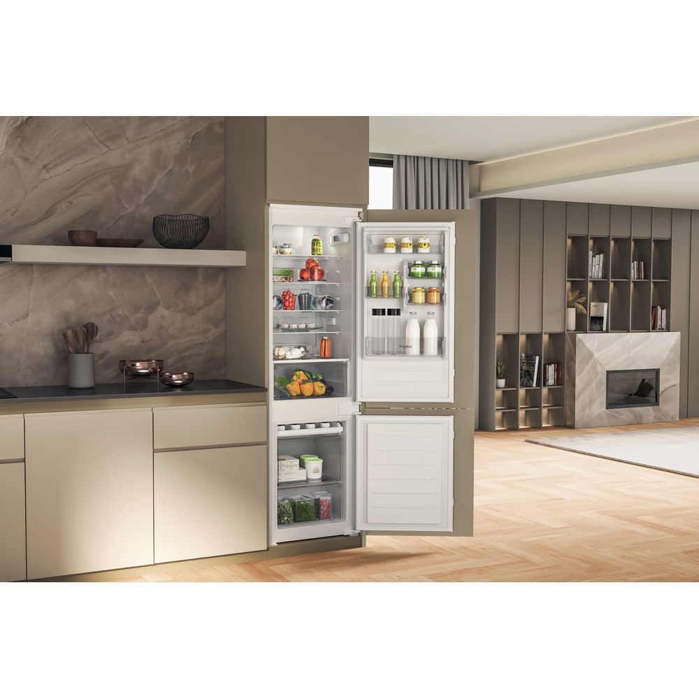 Frigorifero aperto pieno di frutta, verdura e bevande. Cucina con pavimento e armadi in legno. Camino e scaffali sullo sfondo.