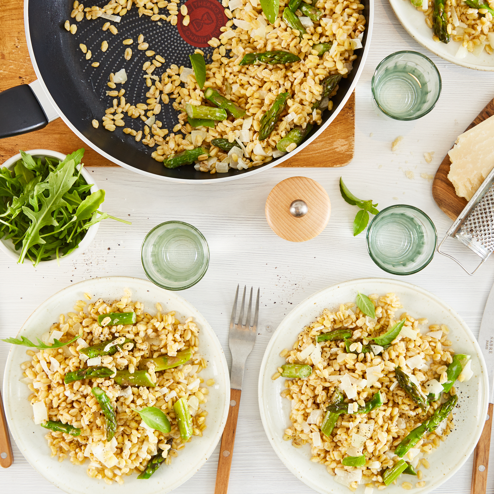 Widok z góry na stół z ugotowanym jęczmieniem i szparagami w patelni i na talerzach, z zielenią i serem.