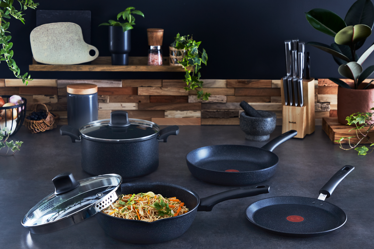 Scena kuchenna z garnkami, patelniami i składnikami. Makaron w patelni. Noże, rośliny i deska do krojenia są również obecne.