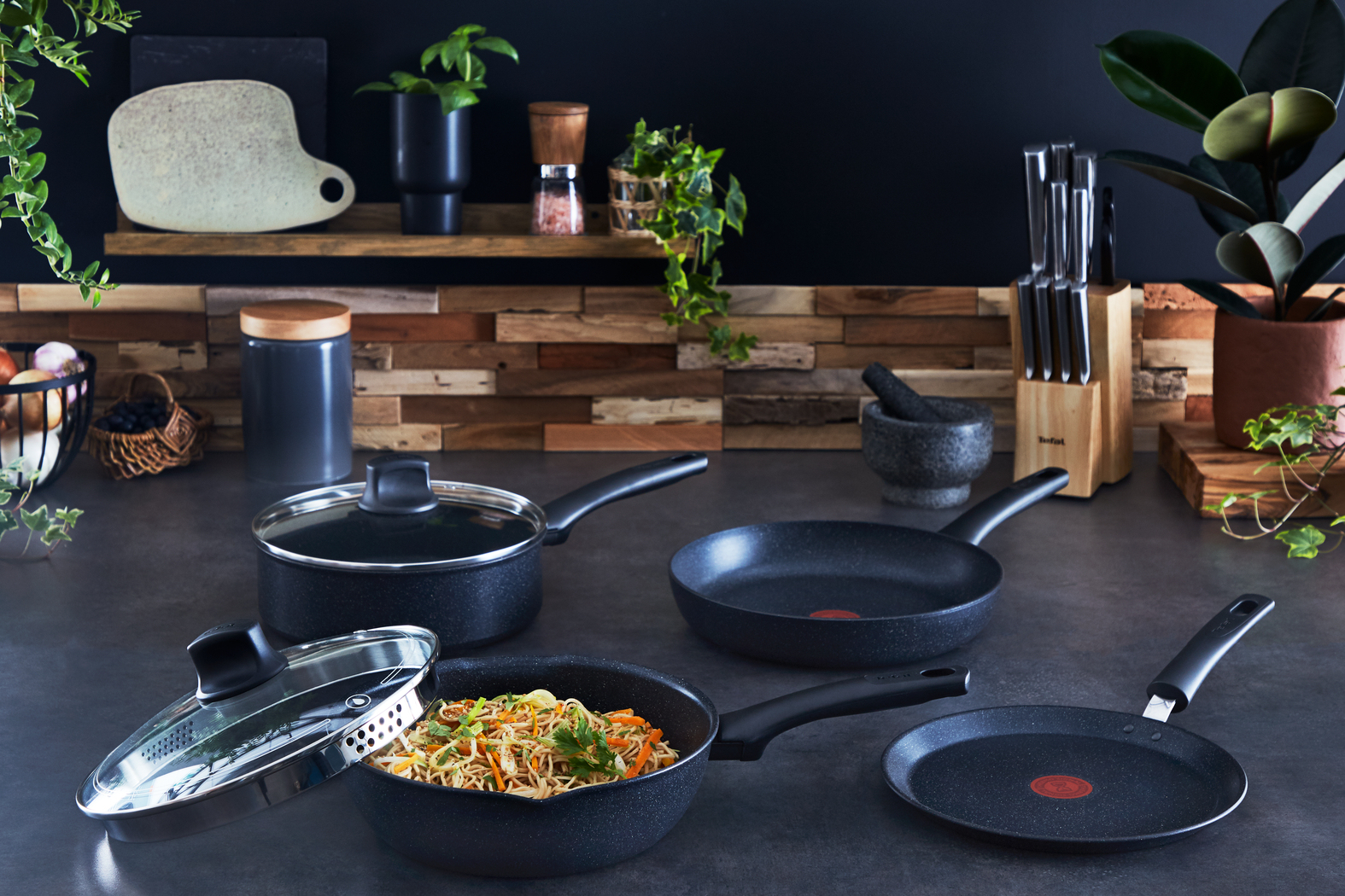 Scena kuchenna z patelniami, jedzeniem i przyborami na ciemnym blacie. Rośliny i półki w tle.