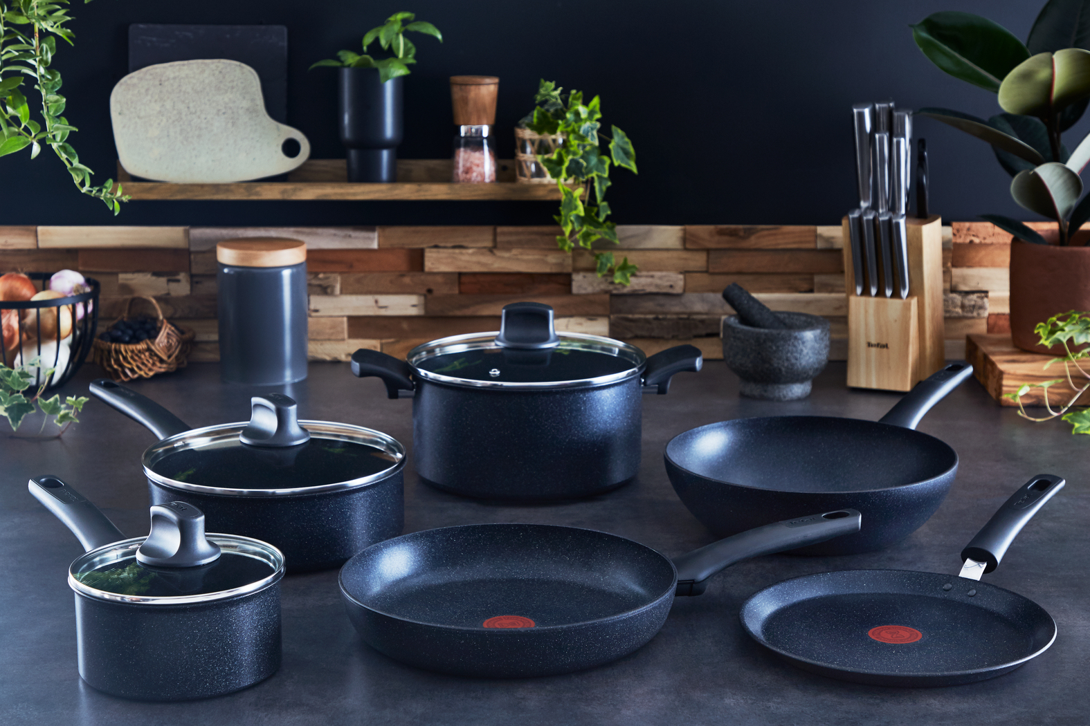 Scena kuchenna z czarnymi naczyniami kuchennymi, roślinami i drewnianymi akcentami. Widoczne patelnie, garnki i noże. Nowoczesne, dobrze oświetlone otoczenie kuchni.