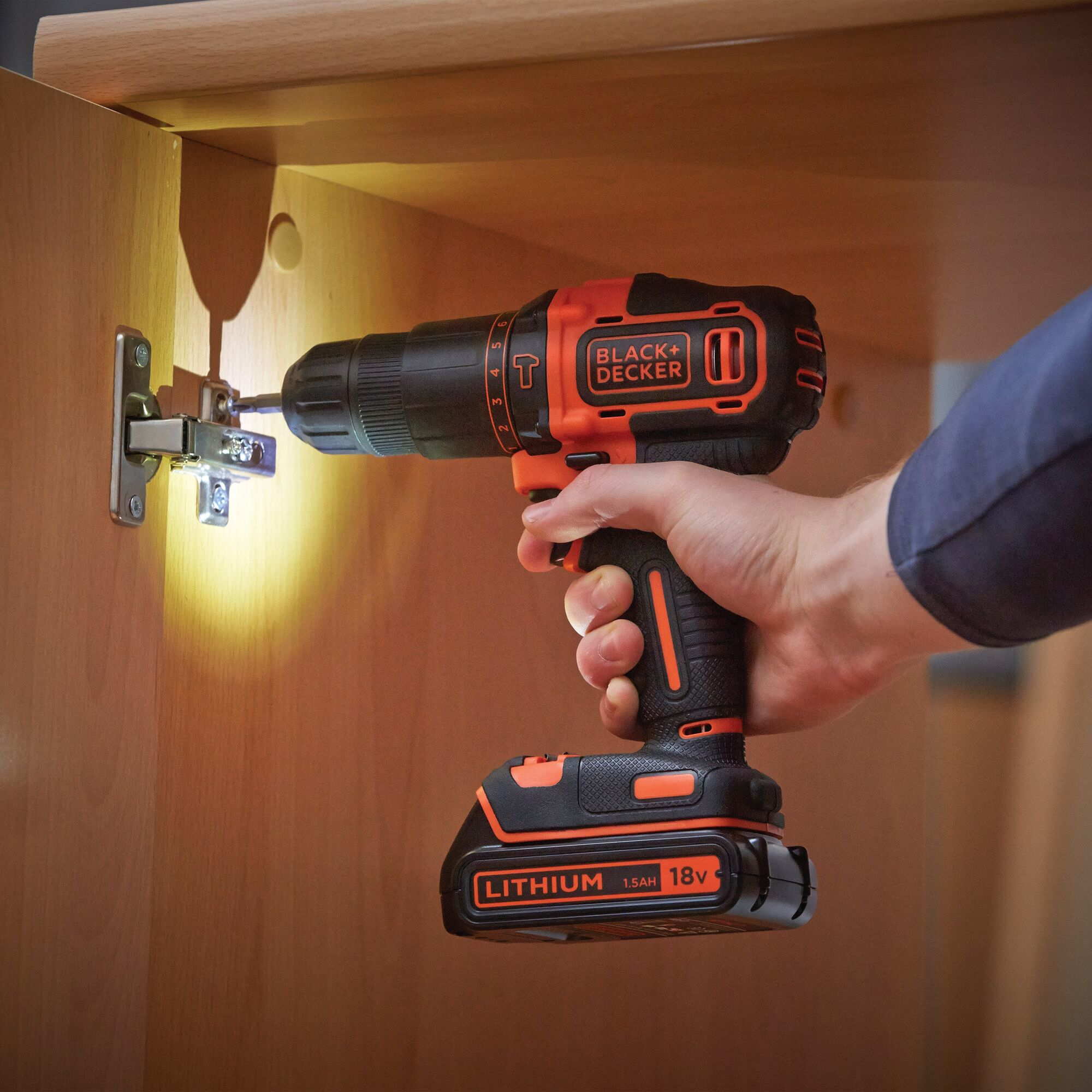 Ręka używająca wiertarki Black+Decker do montażu zawiasu na drewnianej szafce. Scena dobrze oświetlona.