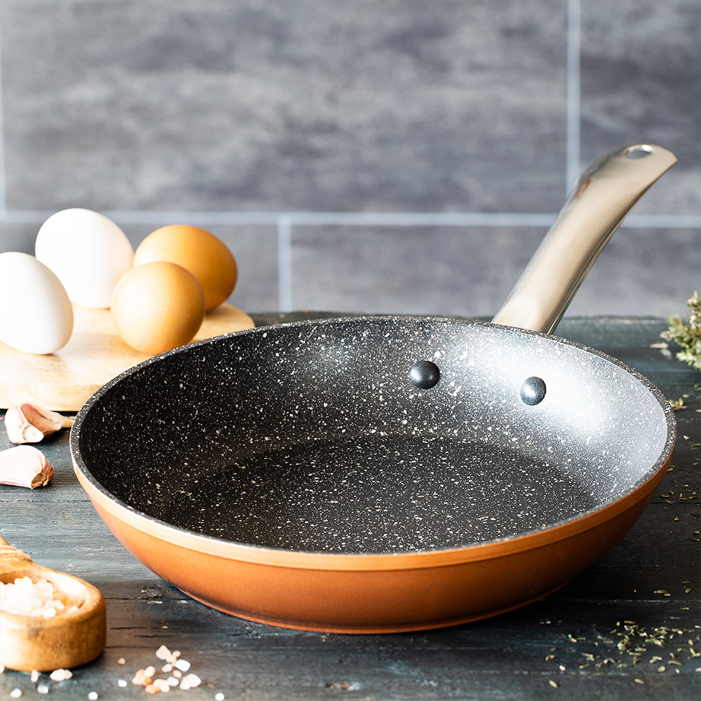 Miedziana patelnia na drewnianym stole. Jajka i czosnek obok patelni. Tłem jest szara, kafelkowa ściana.