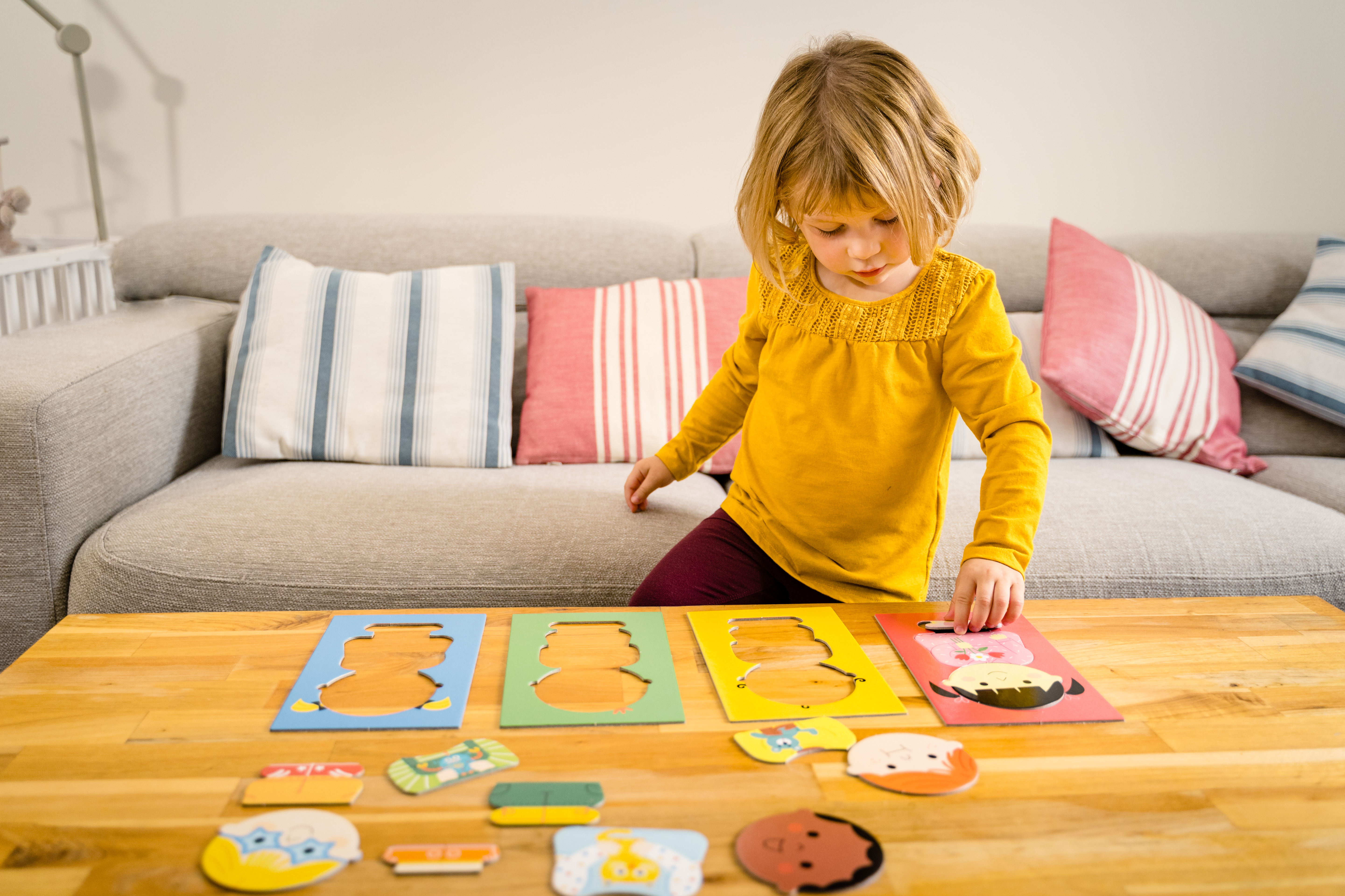 Una niña de amarillo juega con piezas de rompecabezas en una mesa, sofá al fondo.