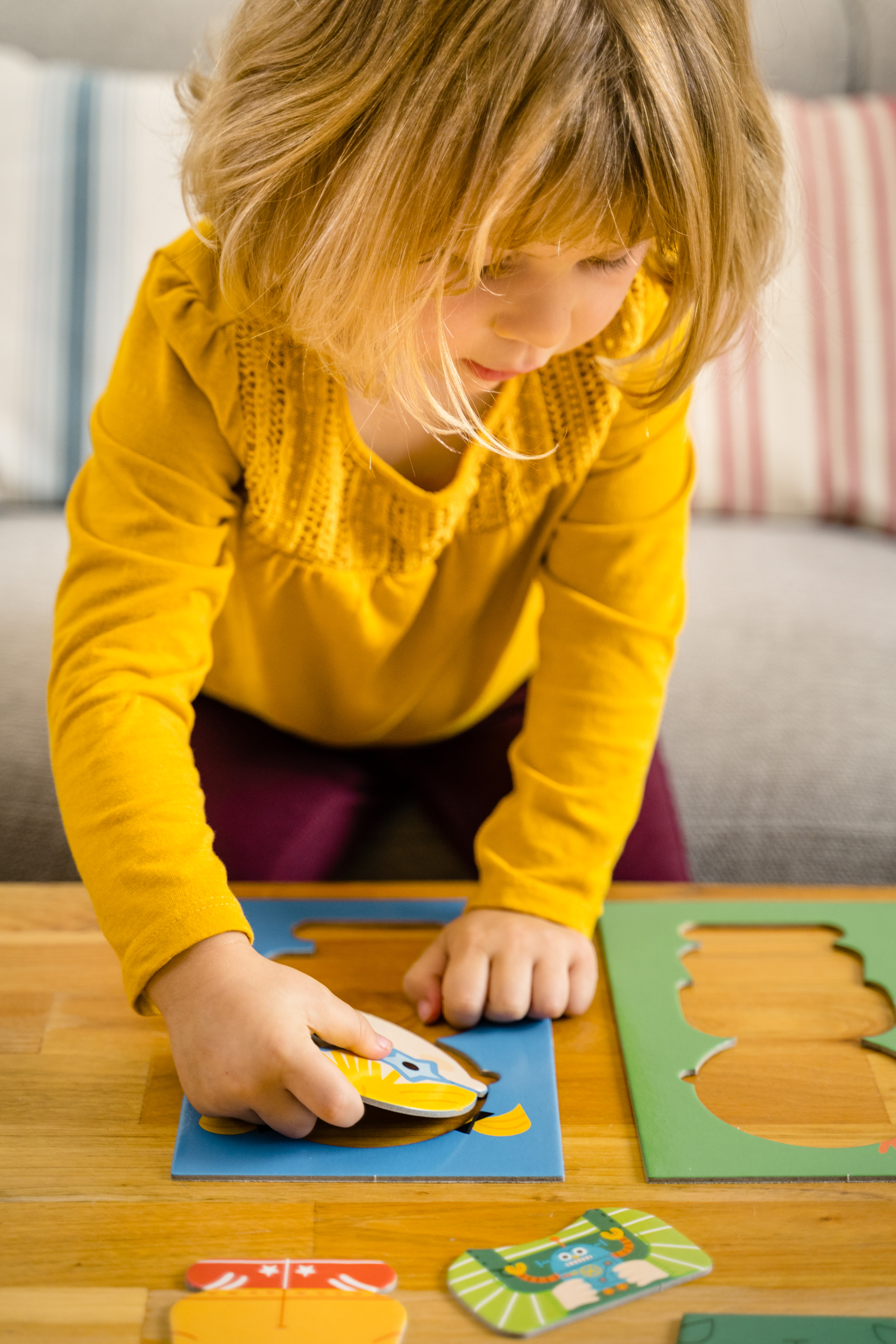 Un niño de amarillo juega con un rompecabezas sobre una mesa de madera. Fondo: sofá con cojín a rayas.