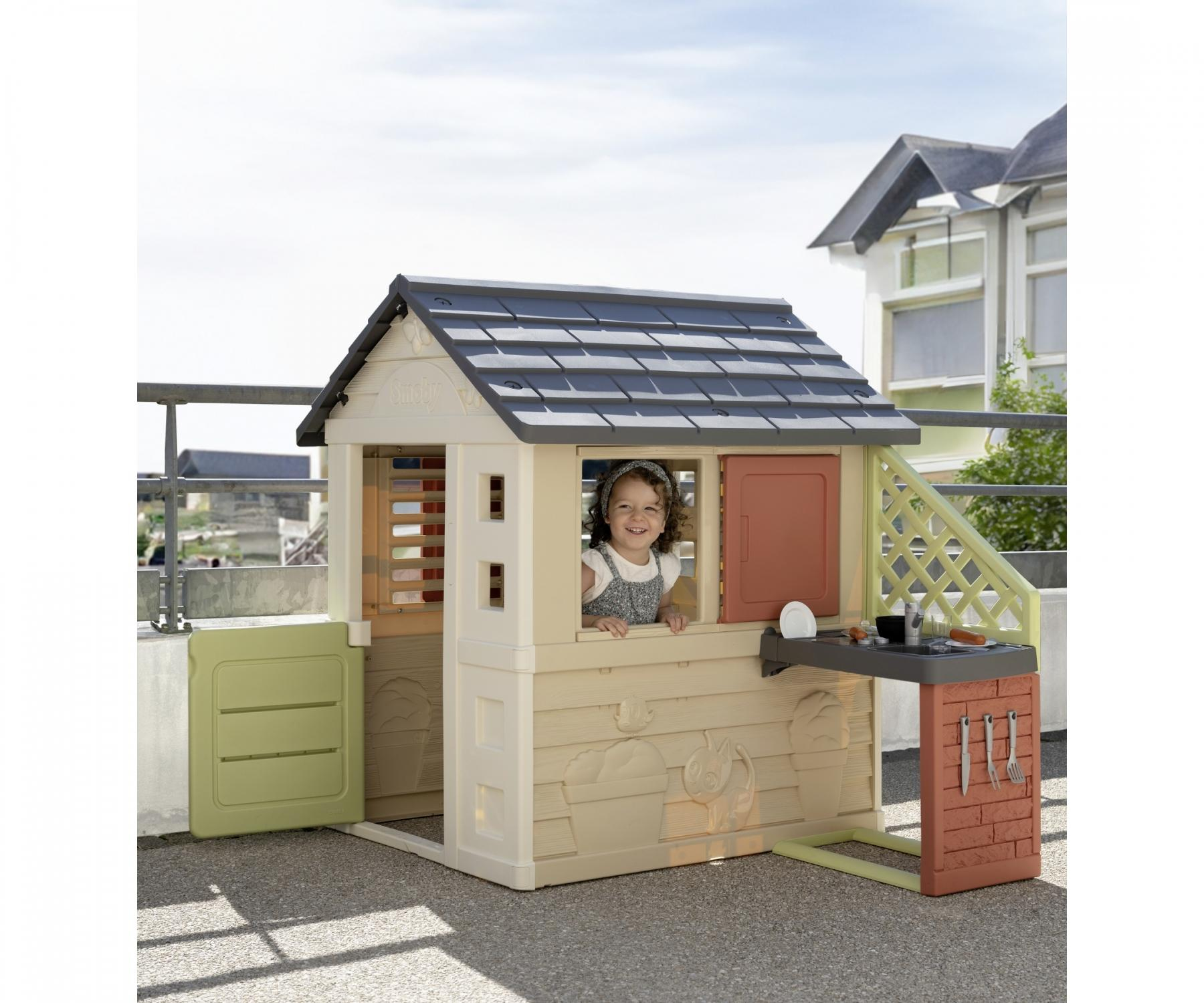 Ein Kind lächelt aus einem Spielhaus. Das Haus ist beige mit grauem Dach. Es gibt eine kleine Küche. Im Hintergrund ist ein Gebäude.