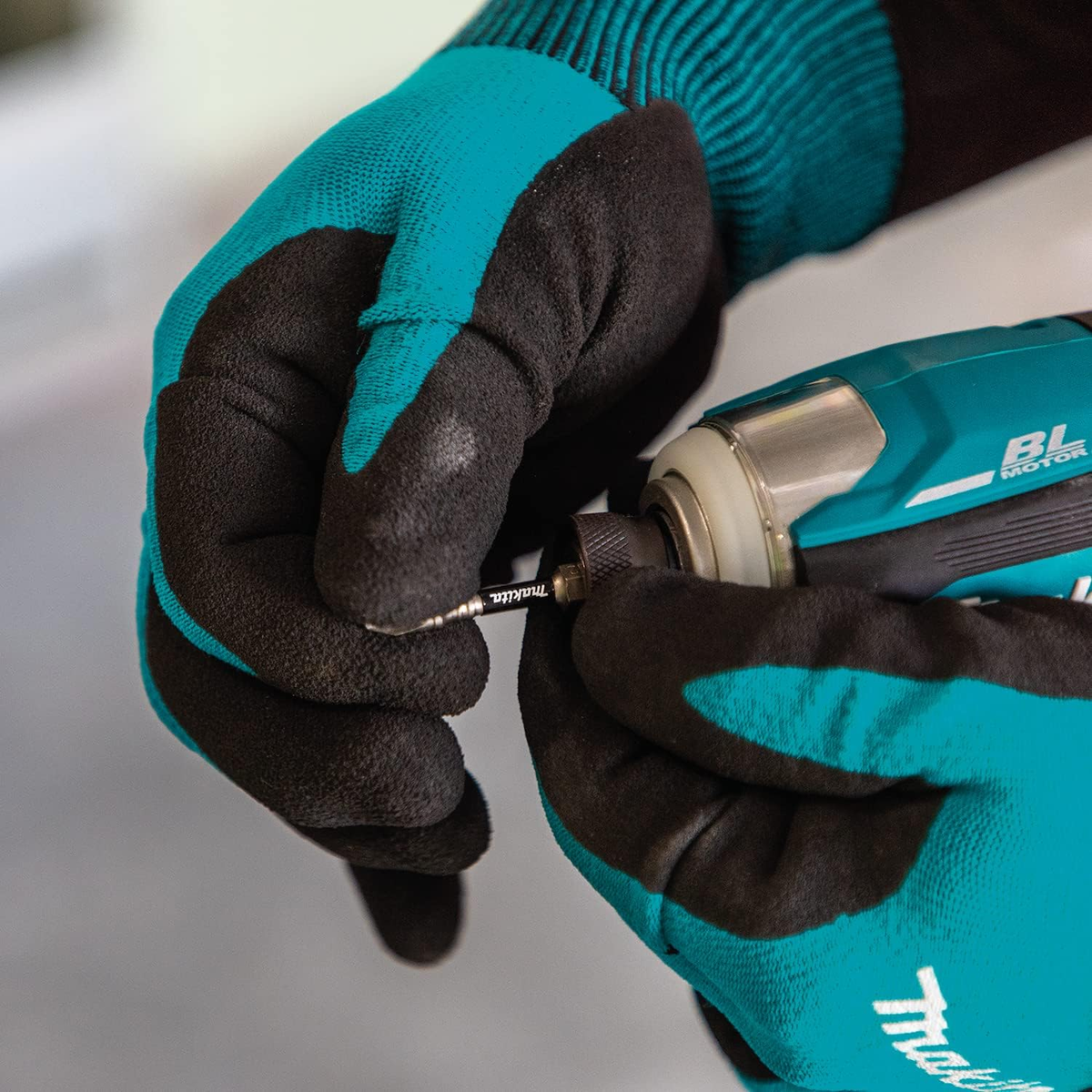 Dłonie w czarno-turkusowych rękawicach trzymają wiertarkę Makita. Wiertło jest wkładane. Tło jest jasne i neutralne.