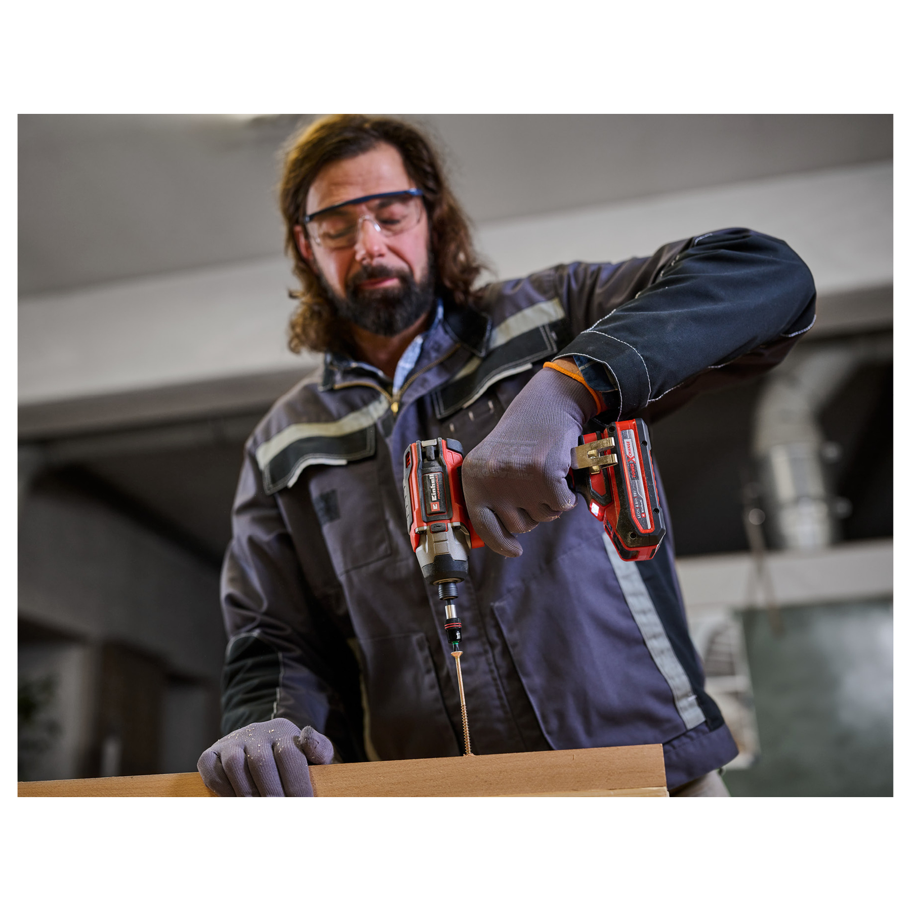 Mężczyzna używa czerwono-czarnej wiertarki do wkręcania w deskę. Nosi okulary ochronne i rękawice robocze.