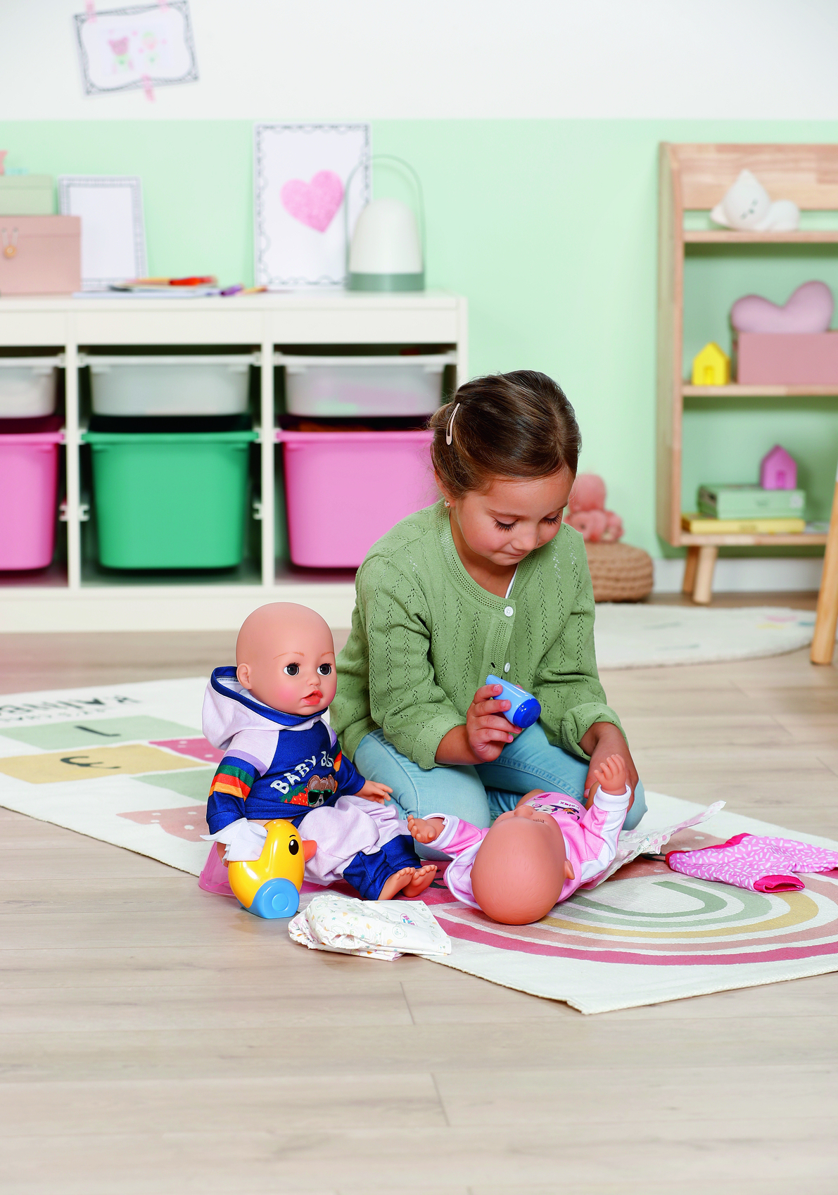 Dziewczynka bawi się lalkami. Jedna lalka leży na macie, druga siedzi. Dziewczynka trzyma niebieską butelkę.