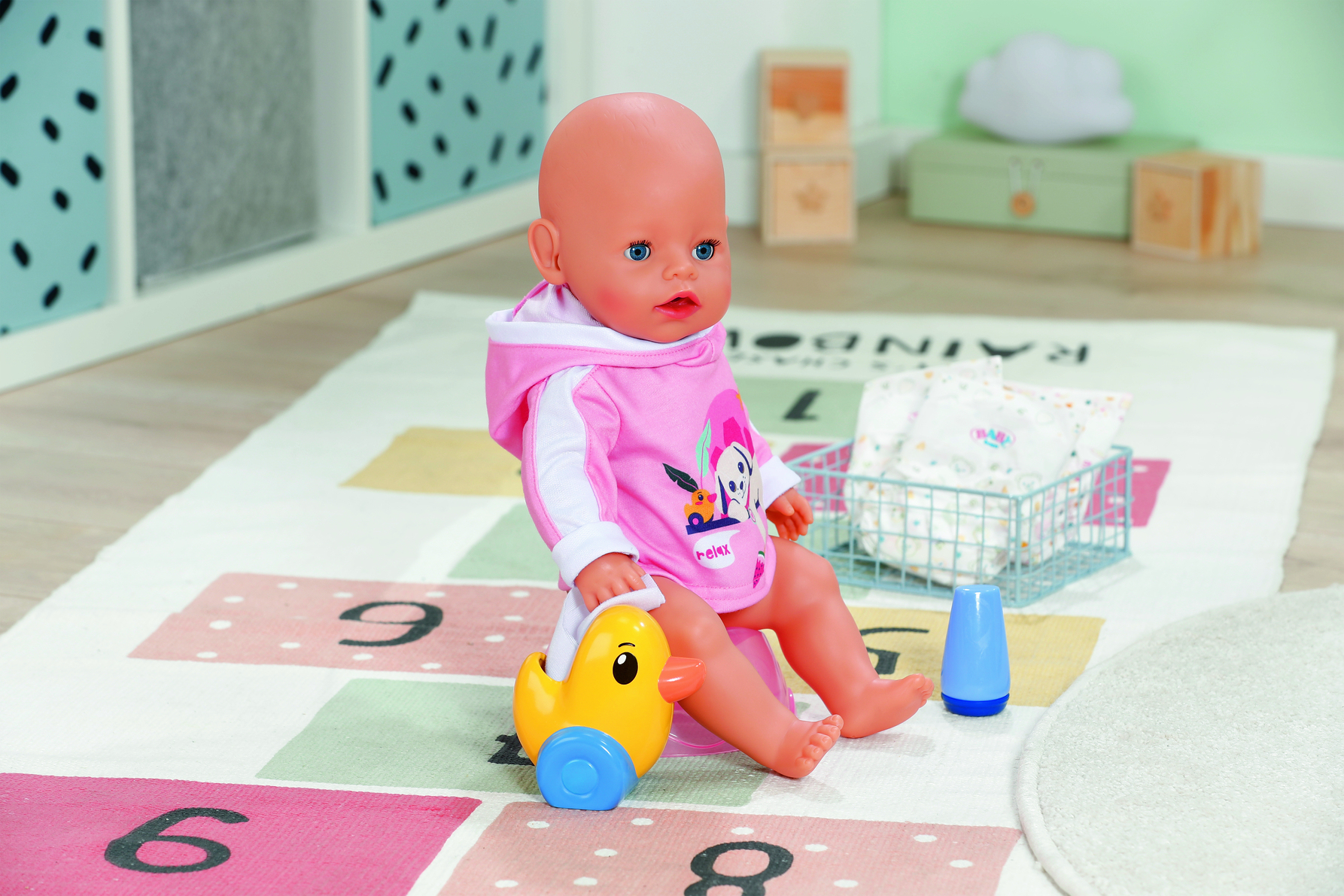 Lalka dla dziecka w różowym stroju, siedząca na kolorowej macie z żółtą zabawką w kształcie kaczki. W pobliżu niebieski kubek i kosz na pieluchy.