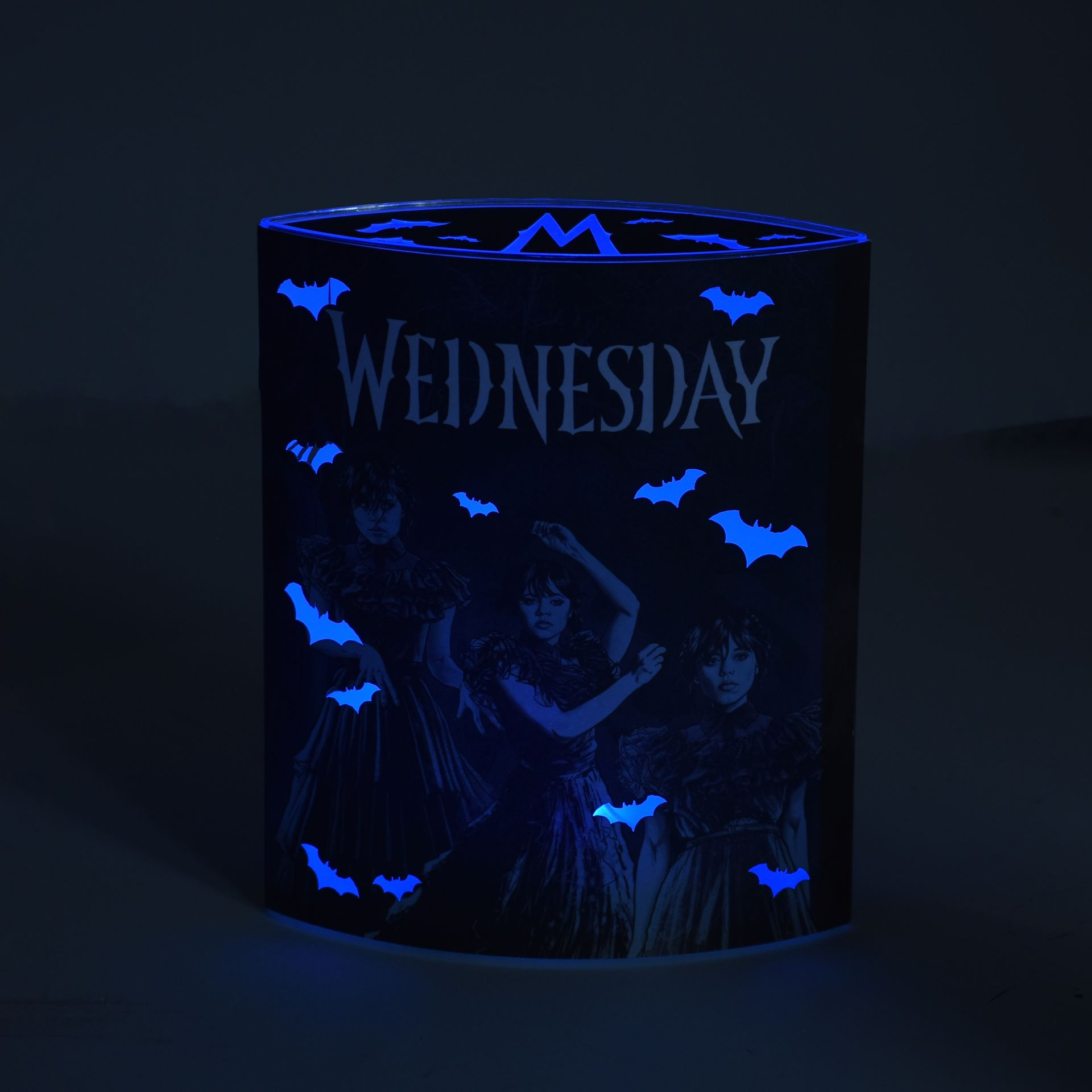 Un cylindre noir avec le mot 'WEDNESDAY', des images de trois femmes, des chauves-souris bleues et un 'M' illuminé.