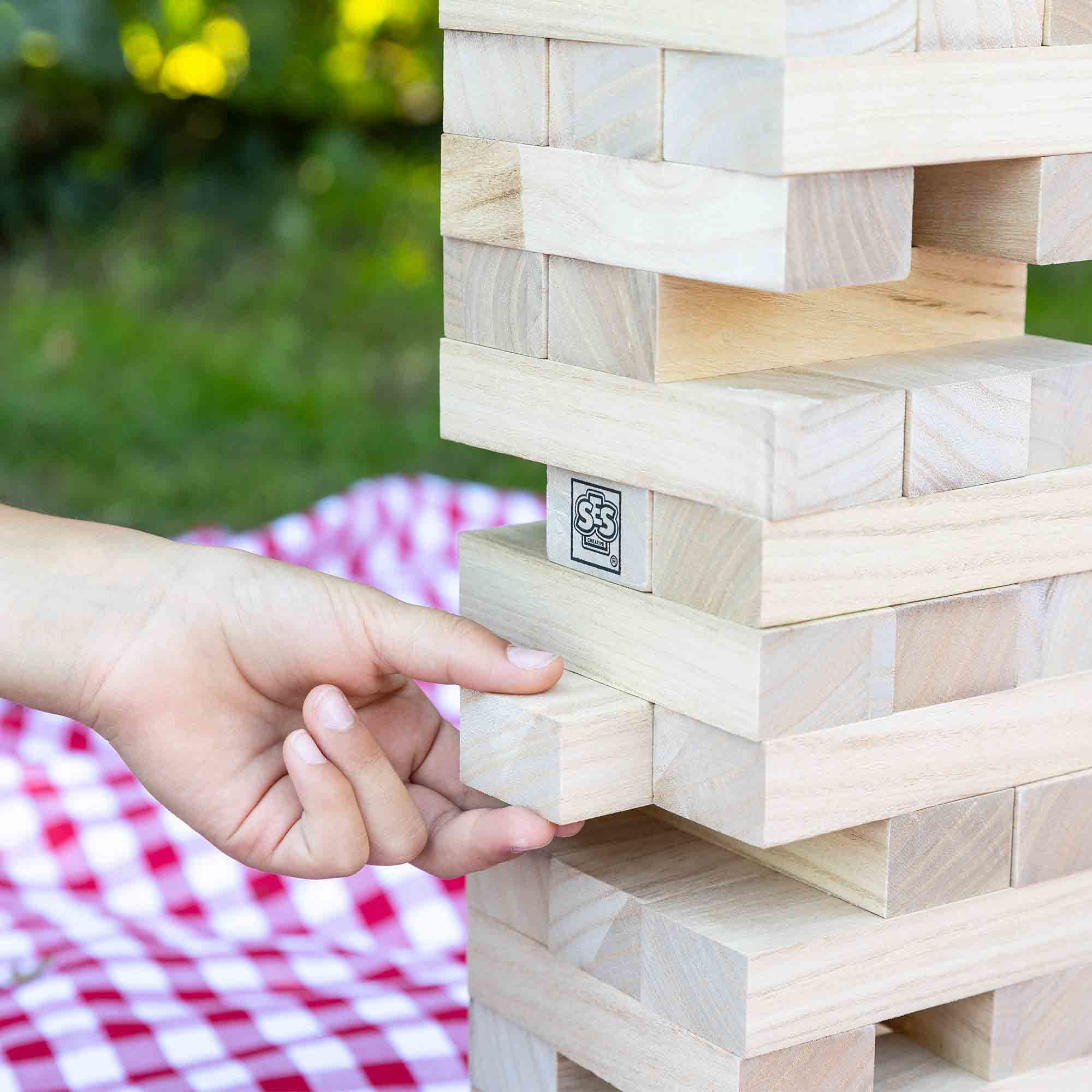 Una mano estrae un blocco di legno da una torre Jenga. Una coperta a scacchi rossi e bianchi è dietro.