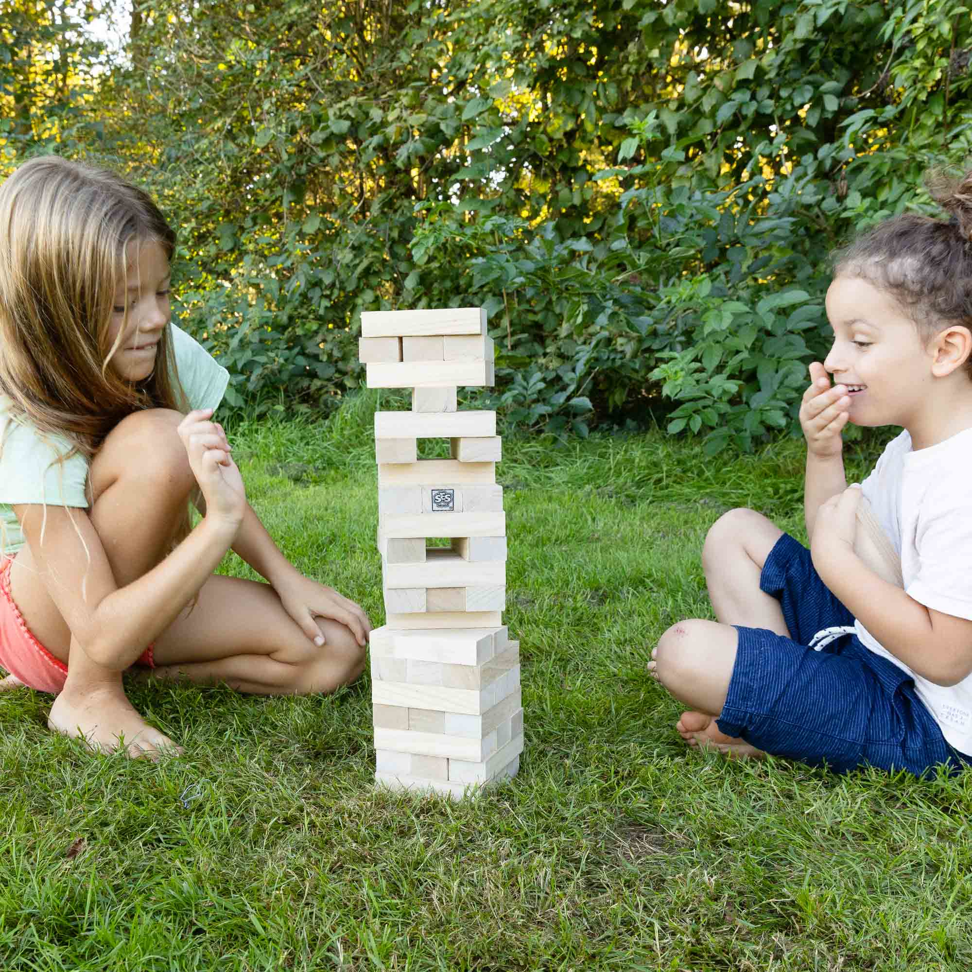 Due bambini giocano a Jenga sull'erba. Uno sta per togliere un blocco, l'altro ride. Cespugli verdi sullo sfondo.