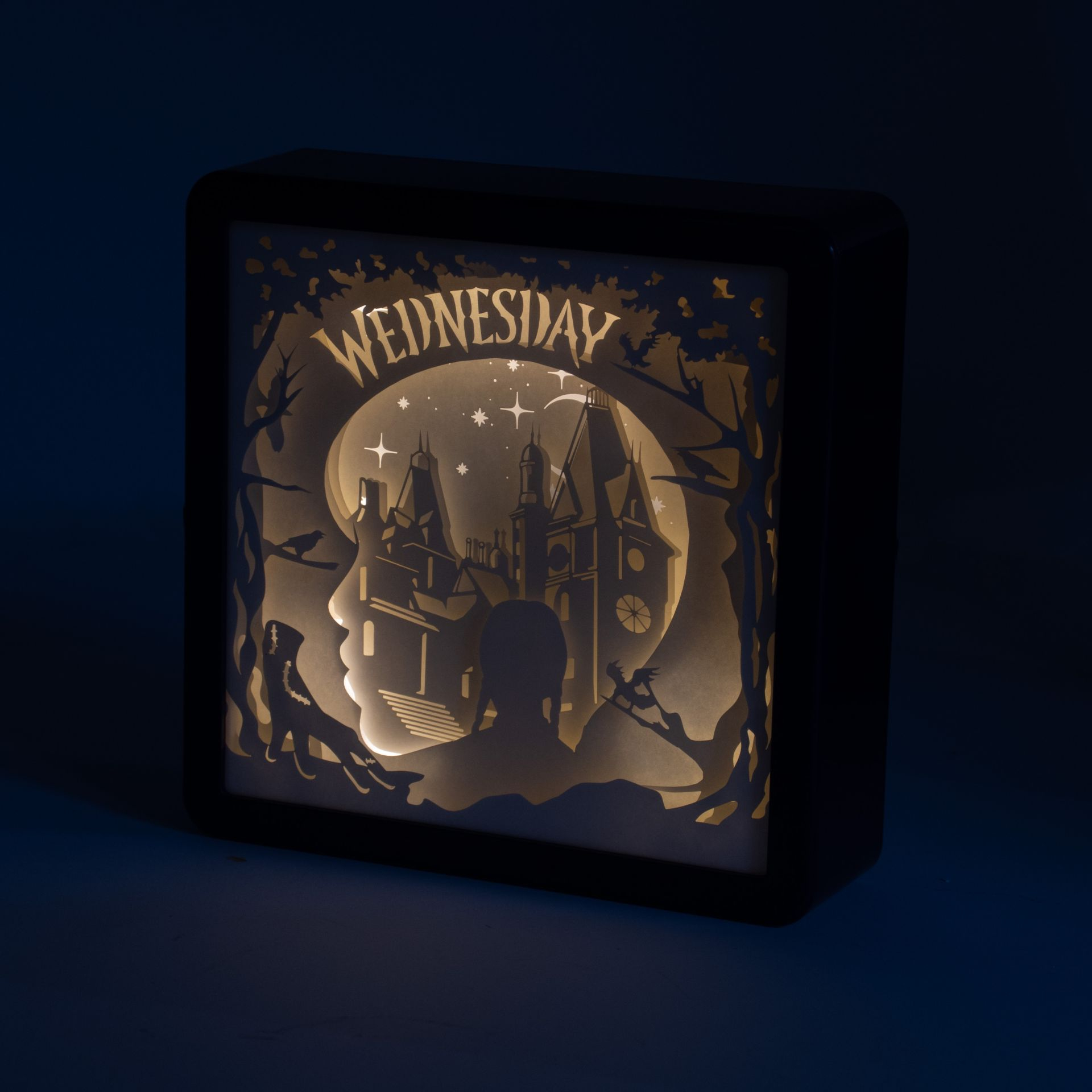 Podświetlana papierowa wycinanka przedstawia zamek, drzewa i sylwetkę Wednesday Addams. Słowo 'Wednesday' jest na górze.