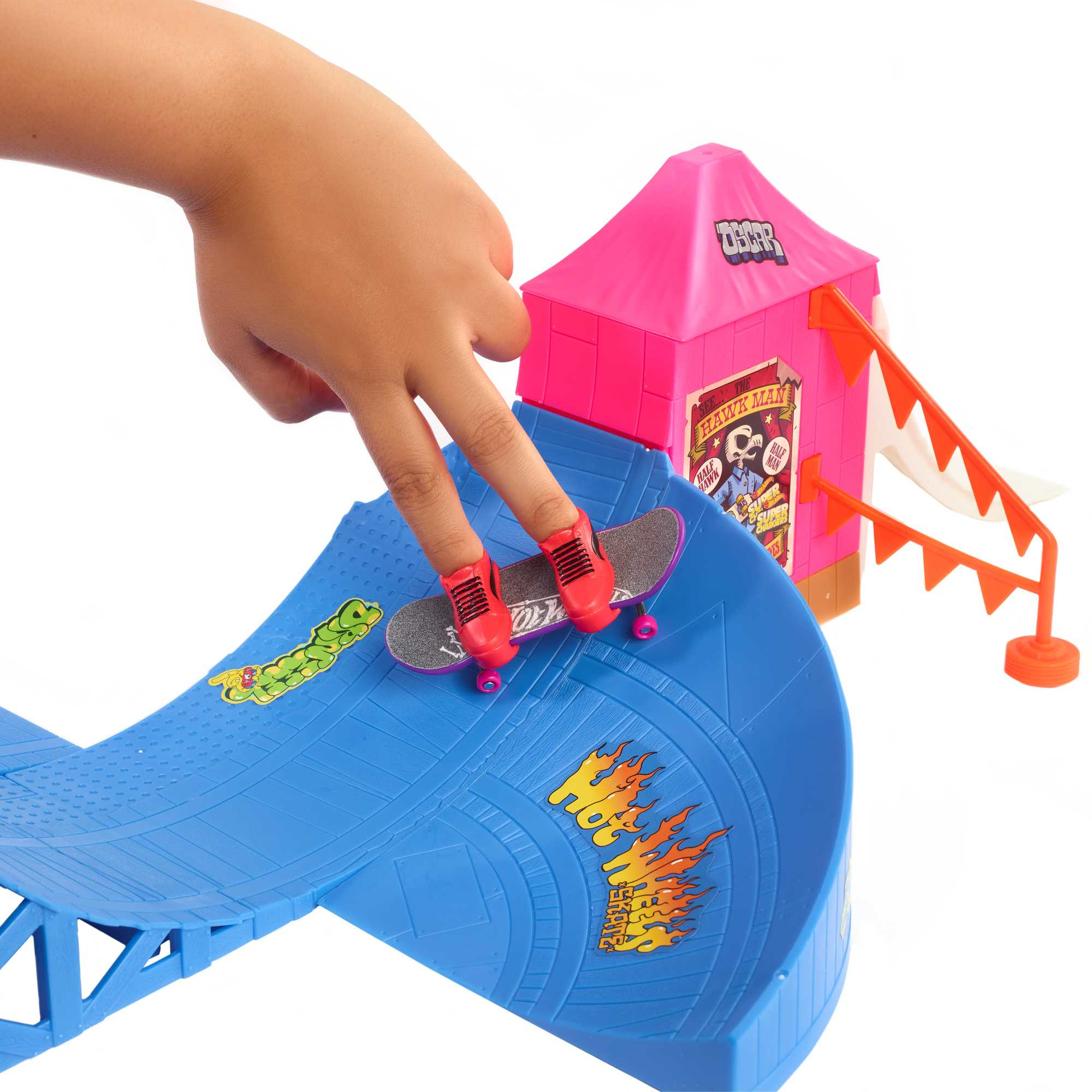 Una mano infantil coloca dedos cubiertos de zapatos rojos sobre un monopatín de juguete en una rampa azul, con una carpa de circo rosa al fondo.