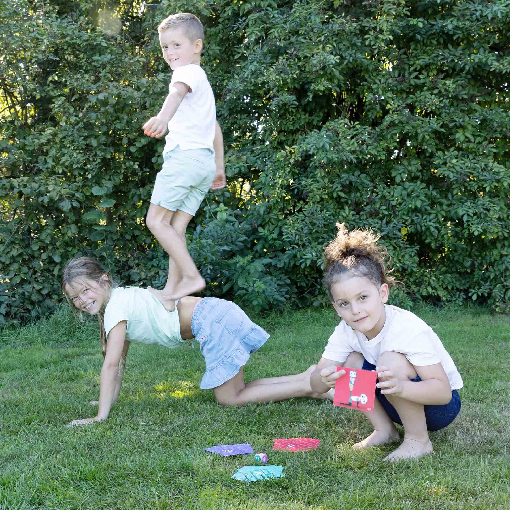 Tre bambini che giocano sull'erba. Uno salta, un altro è a terra e l'ultimo tiene una carta. Sfondo verde.