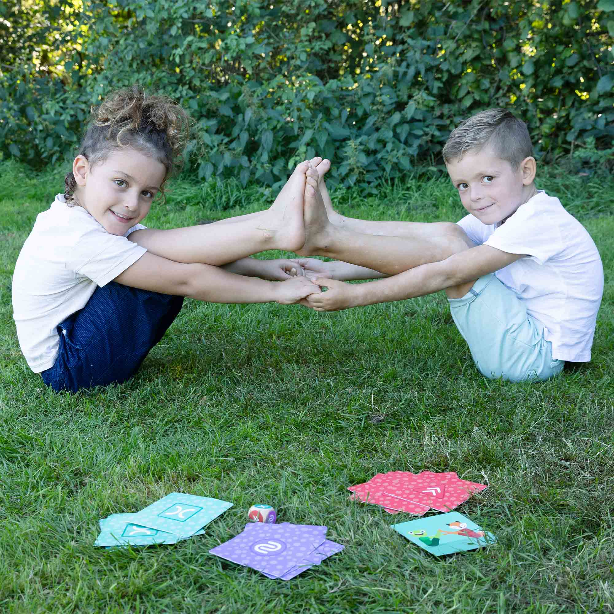 Due bambini si tengono per mano e per i piedi, sorridendo. Carte e un dado sull'erba.