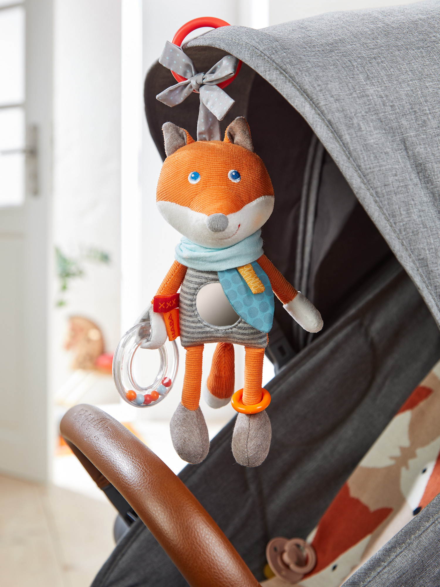 Ein Fuchspielzeug hängt an einem grauen Kinderwagen. Der Fuchs hat orangefarbenes Fell, blaue Augen und einen blauen Schal. Eine durchsichtige Rassel ist in seiner Hand.
