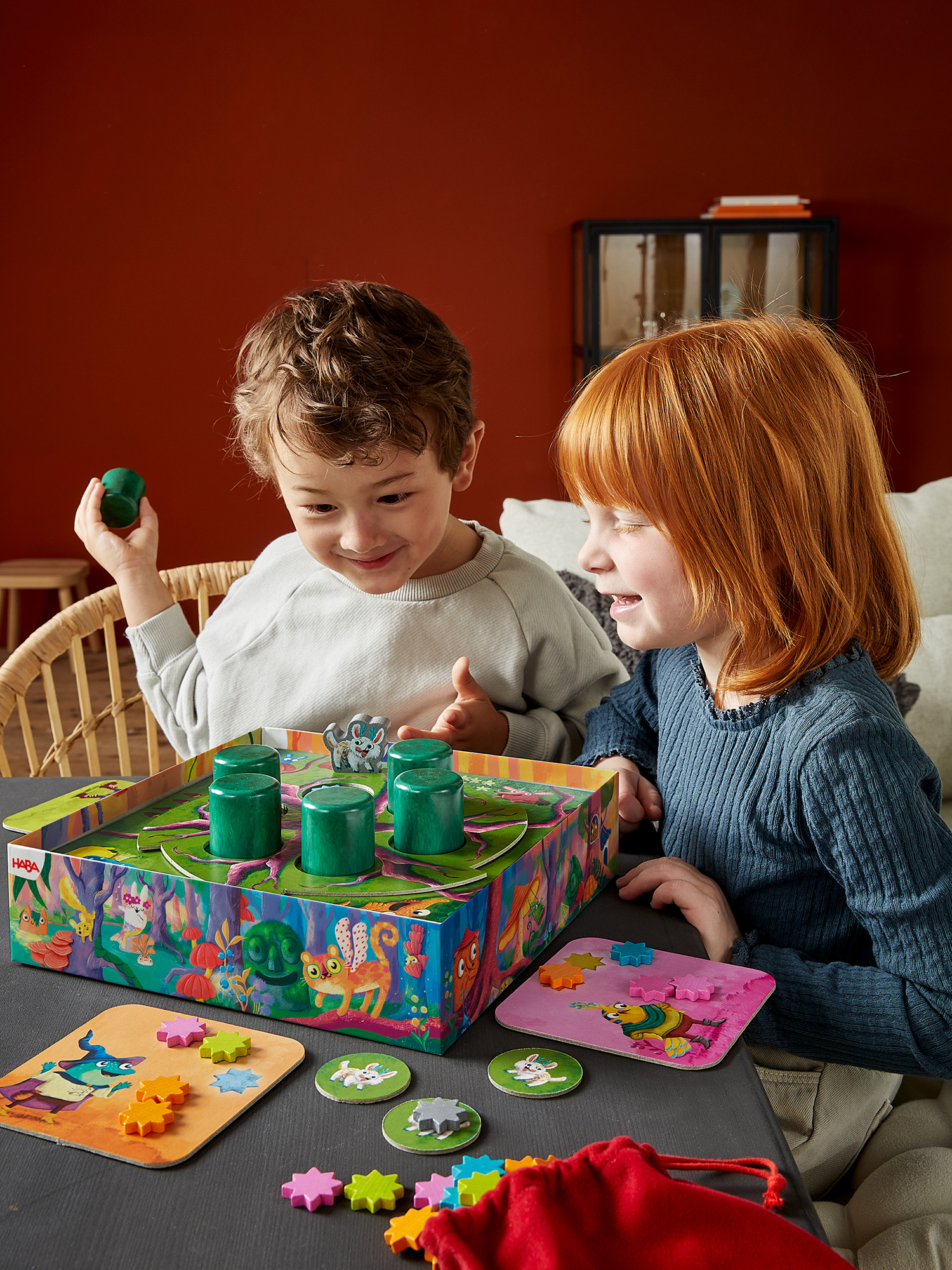 Due bambini giocano a un gioco da tavolo. Il ragazzo tiene in mano un pezzo verde. La ragazza sorride. Un tabellone colorato è su un tavolo scuro.