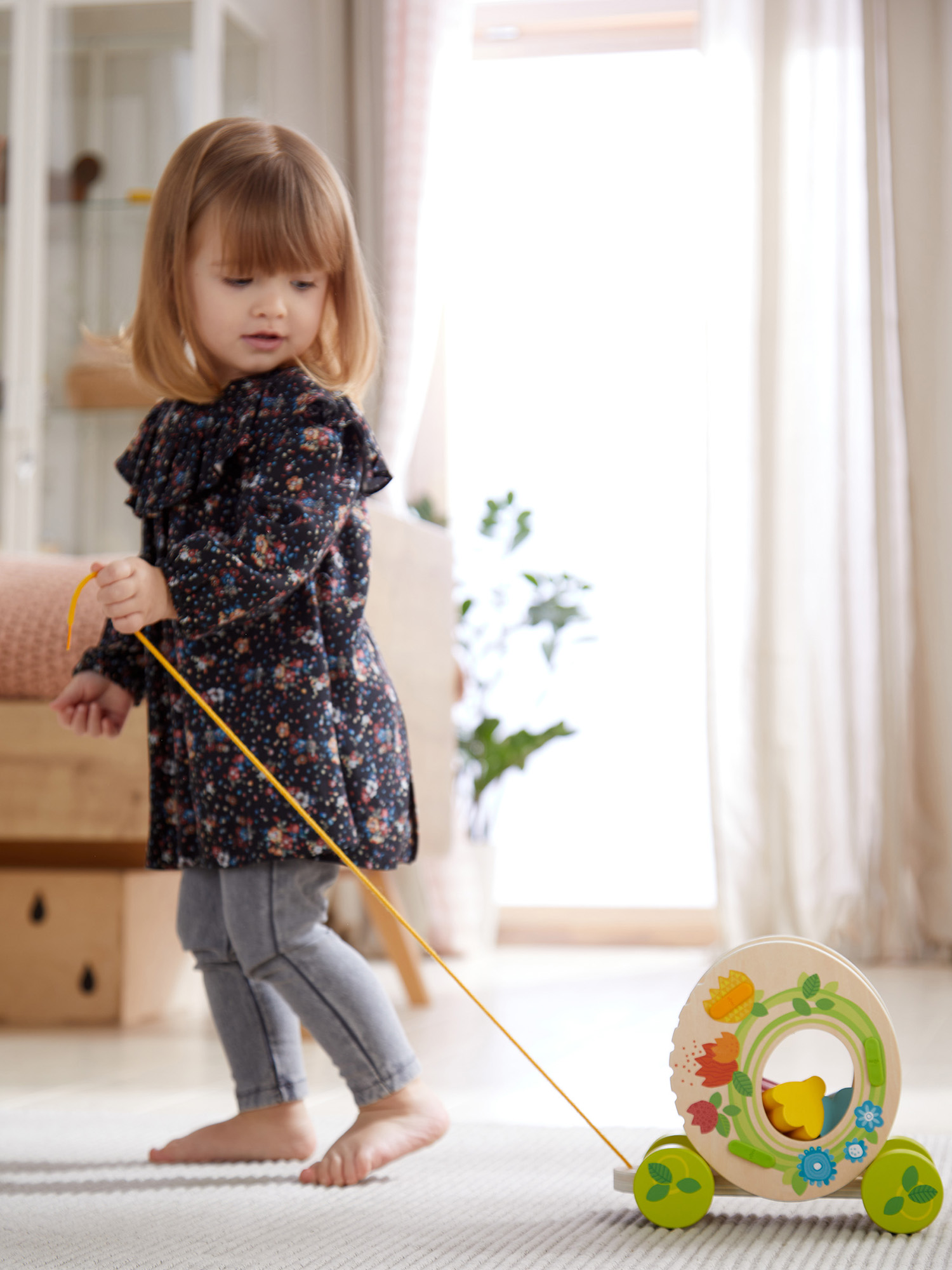 Ein junges Mädchen zieht ein Holzspielzeug. Das Spielzeug ist ein Rad mit Blumendesigns. Sie trägt ein Blumenkleid und Jeans.
