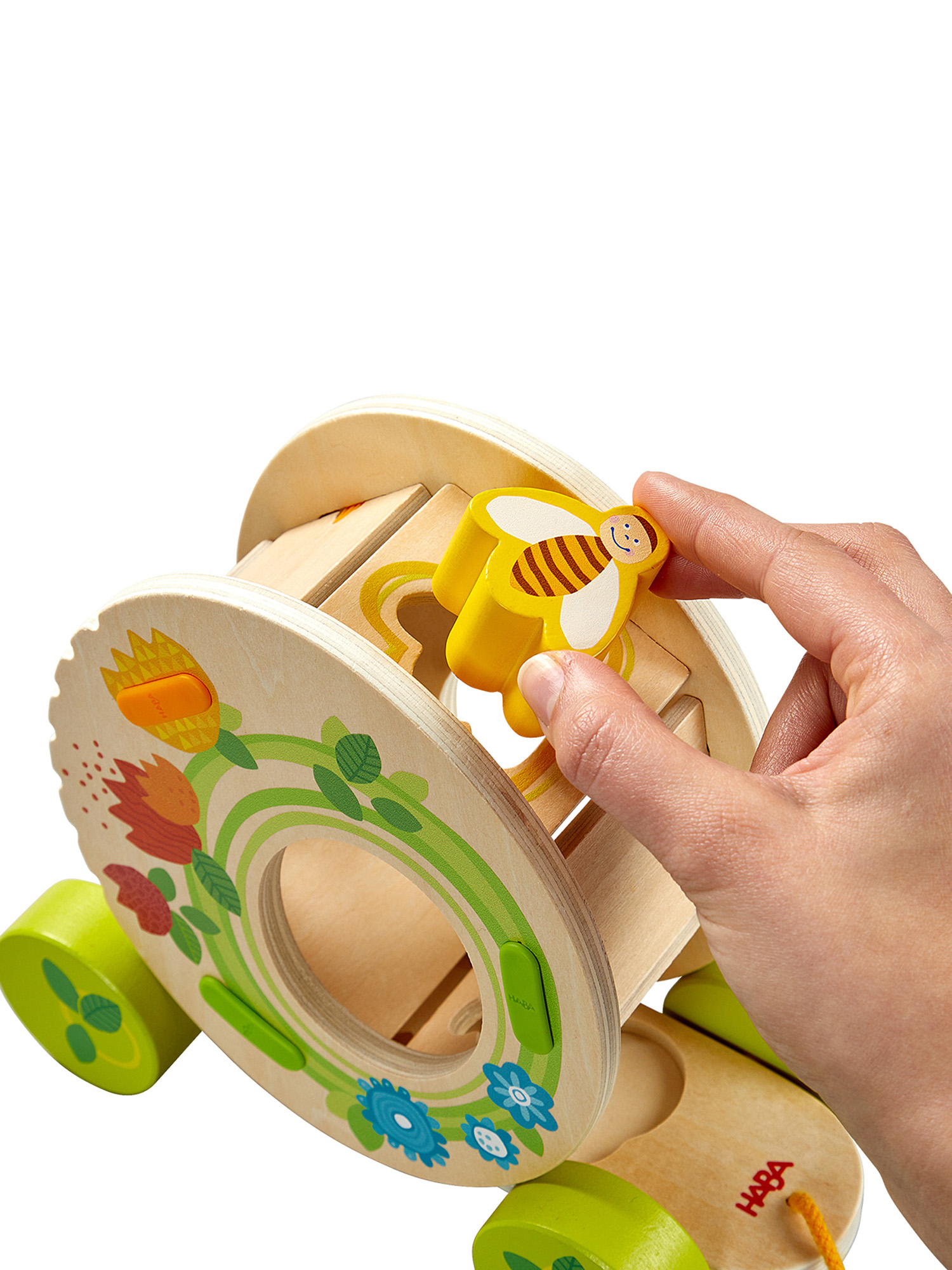 Eine Hand berührt ein Holzspielzeug mit einem bienenförmigen Teil. Das Spielzeug hat Räder und Blumendesigns. Der Hintergrund ist weiß.