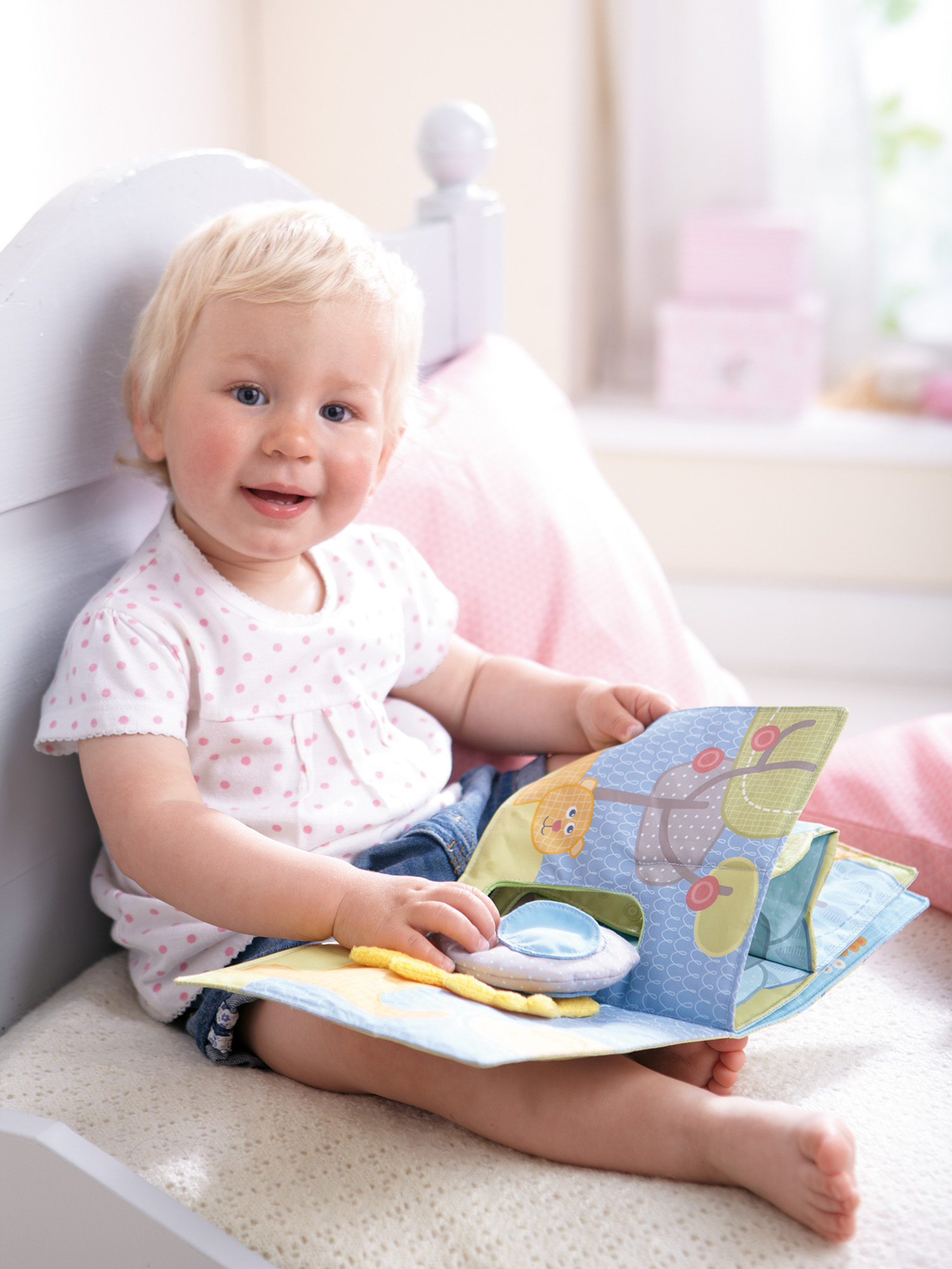 Ein lächelndes Baby mit blonden Haaren hält ein Buch. Sie trägt ein weißes Polka-Dot-Shirt und blaue Jeansshorts. Das Buch ist aufgeschlagen.