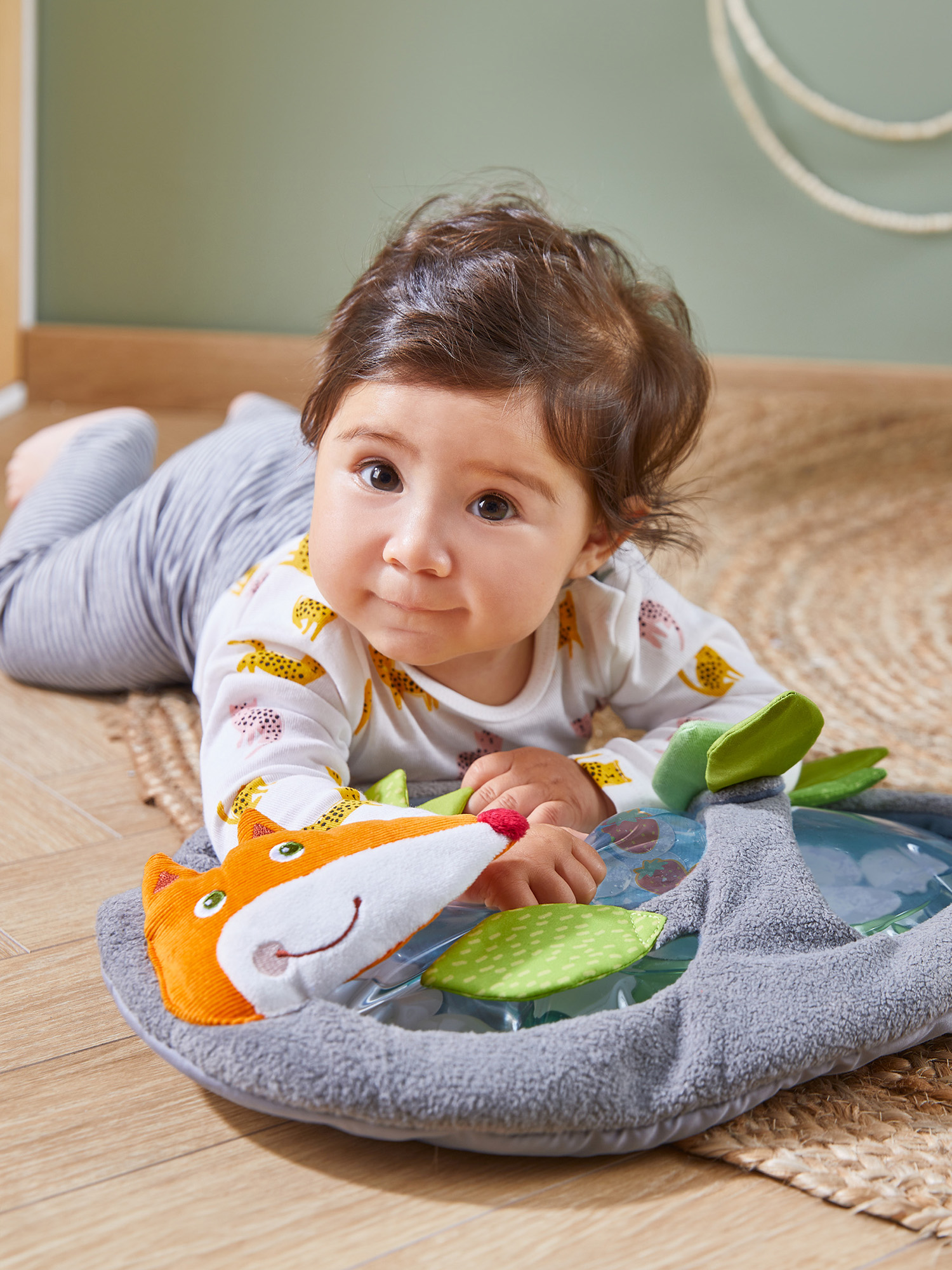 Ein Baby liegt auf einem Teppich und schaut in die Kamera. Es spielt mit einem fuchsähnlichen Spielzeug. Der Hintergrund hat eine grüne Wand und ein Seil.