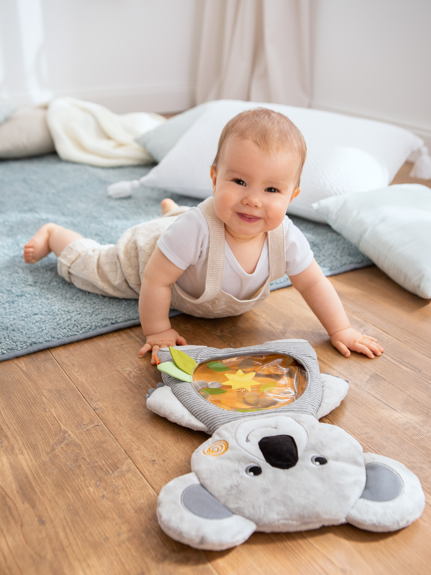 Ein Baby liegt auf einem Holzboden und lächelt. Ein Koala-Spielzeug liegt vor dem Baby. Kissen und ein Teppich sind im Hintergrund.