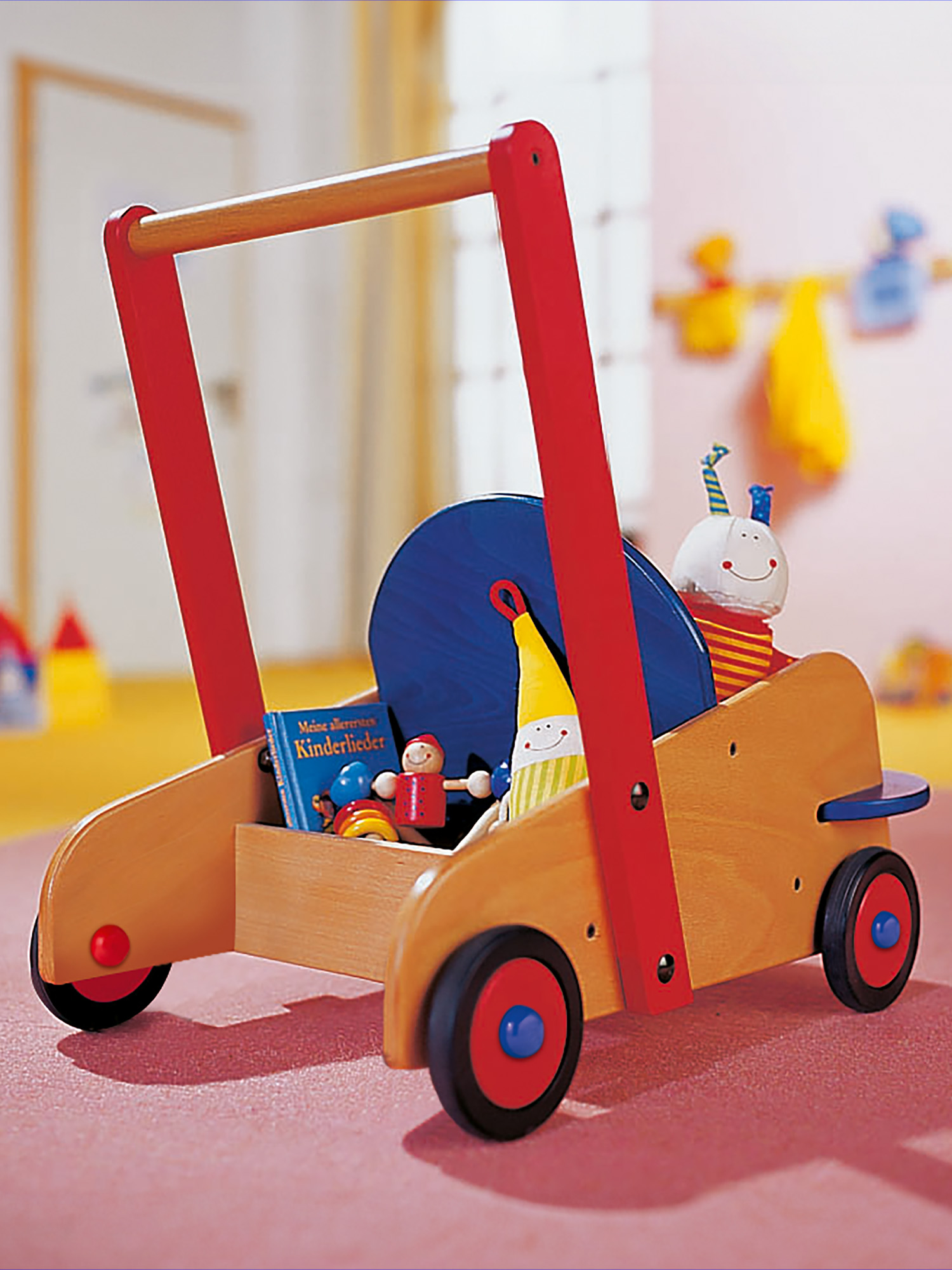 Ein hölzerner Spielzeug-Walker mit roten und blauen Details. Es hat ein blaues Rad, ein Buch und verschiedene Spielzeuge im Inneren. Der Hintergrund ist eine rosa Wand.