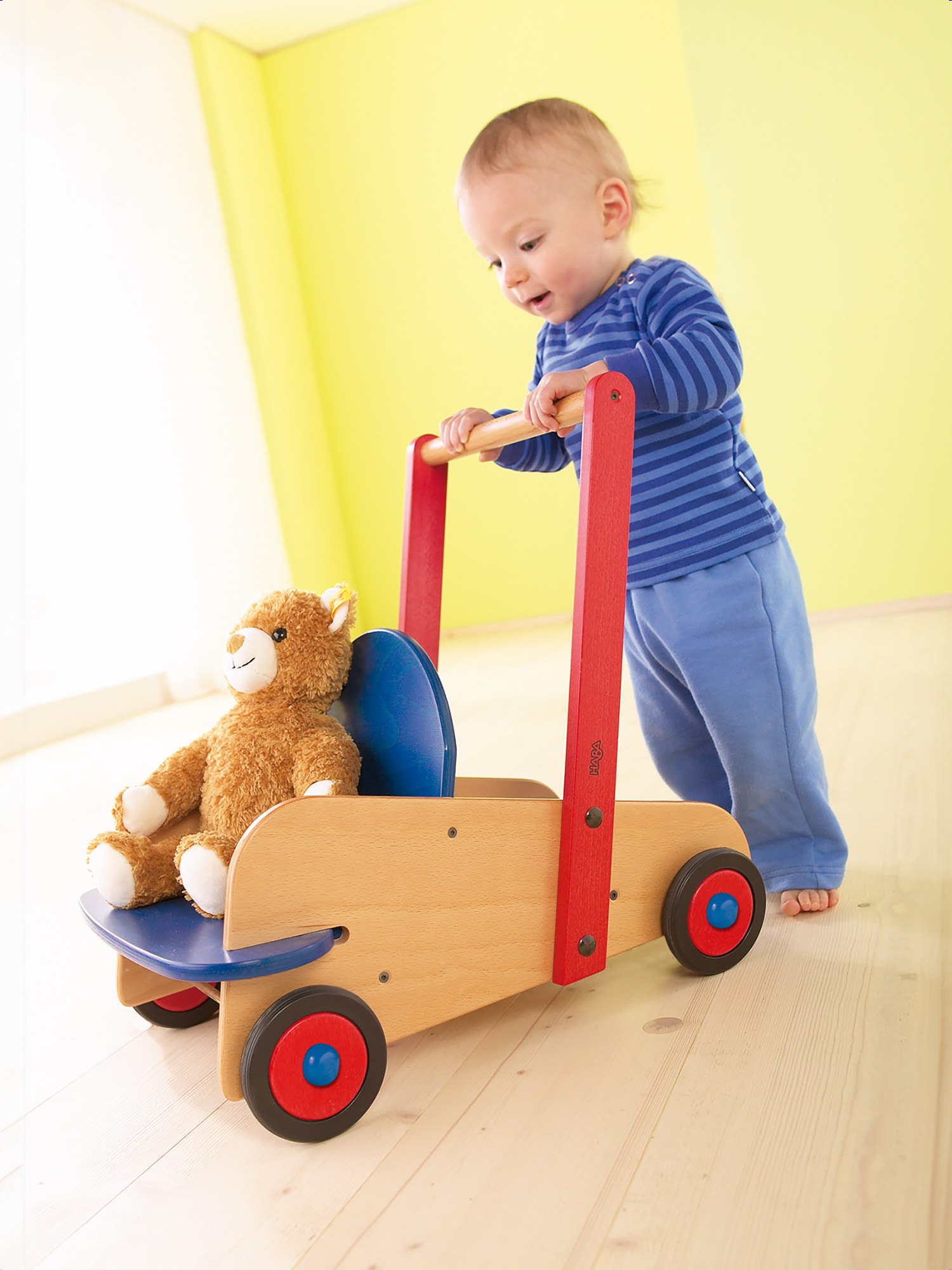 Ein Baby schiebt einen Holzwalker mit einem Teddybären. Der Walker hat rote und blaue Akzente. Das Baby trägt Blau.