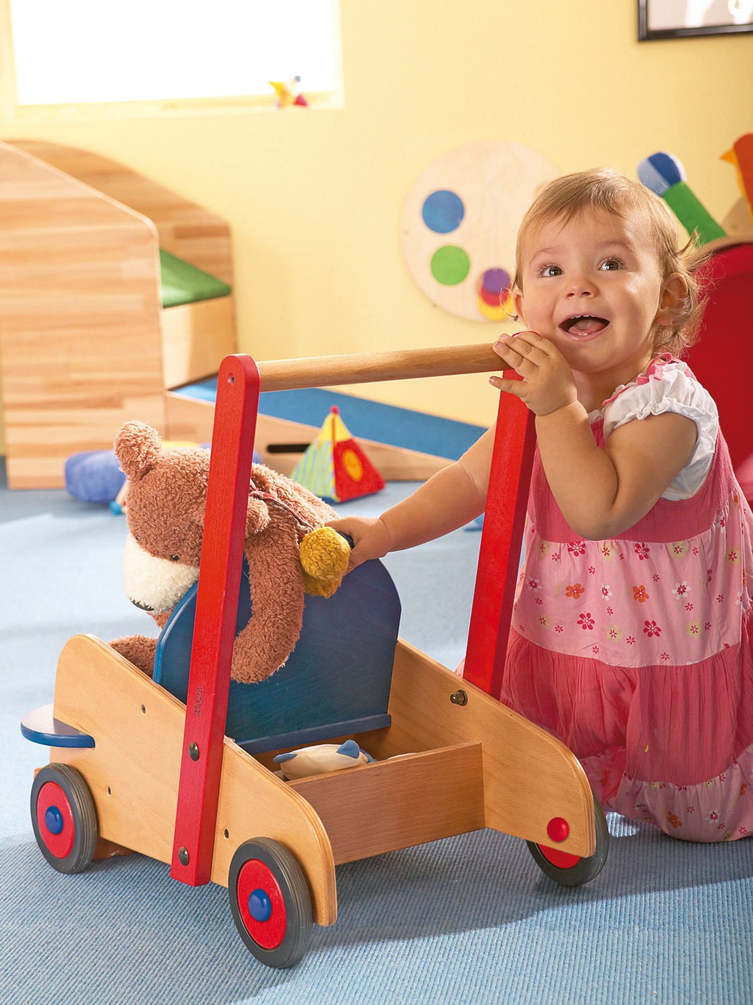 Ein Kleinkind in einem rosa Kleid lächelt, während es einen hölzernen Spielzeugwagen mit einem Teddybären hält. Der Wagen hat rote und blaue Akzente.