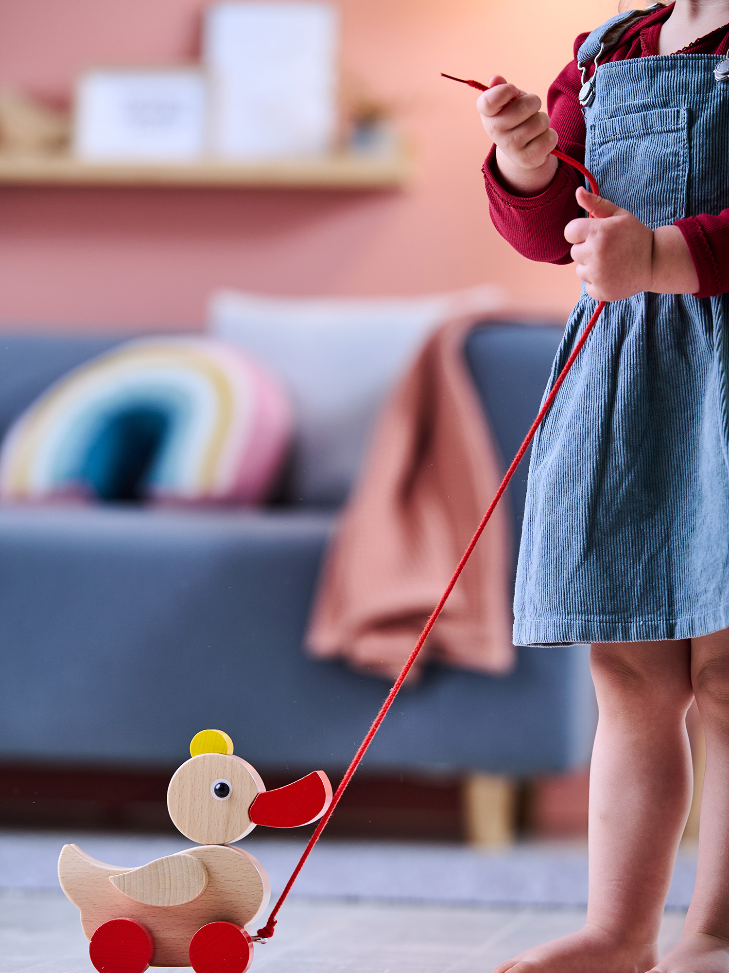 Ein Kind hält eine rote Schnur, die an einer hölzernen Nachzieh-Ente befestigt ist. Das Kind trägt ein blaues Kleid. Der Hintergrund zeigt ein Sofa.