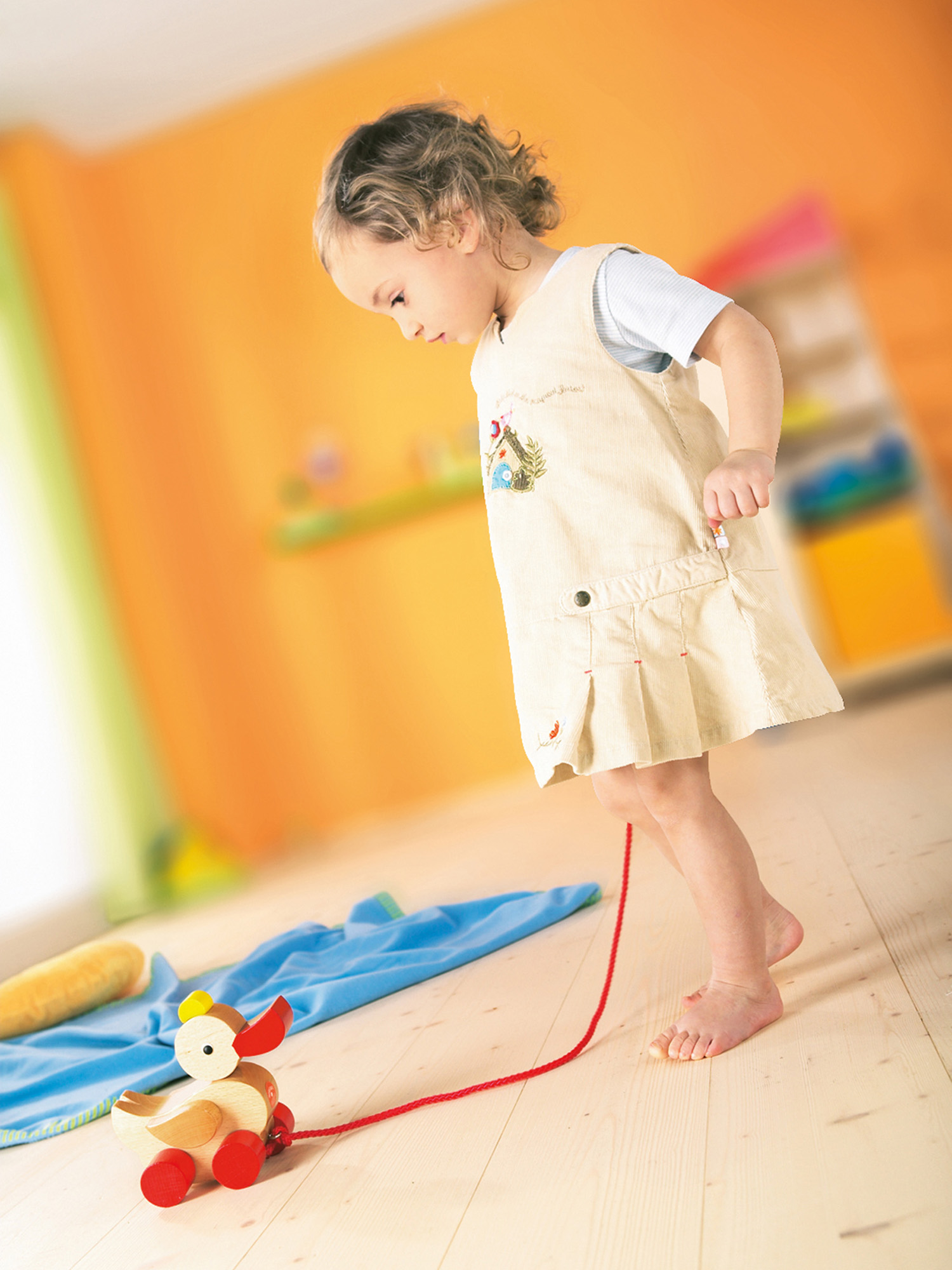 Ein junges Mädchen in einem Kleid zieht eine Spielzeugente an einer Schnur. Der Hintergrund ist eine orangefarbene Wand und ein Holzboden.