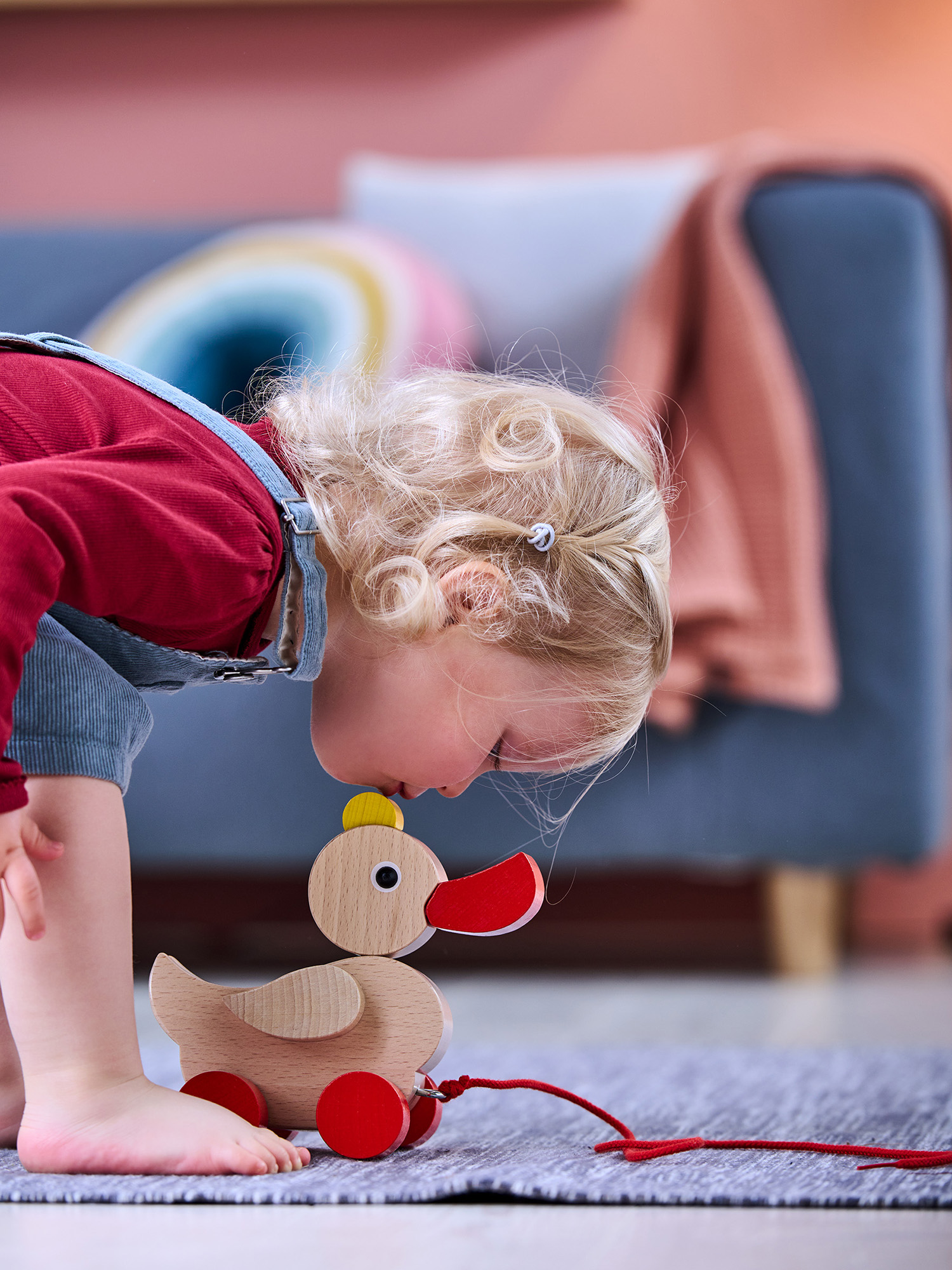 Ein Kind betrachtet ein hölzernes Entenspielzeug auf einem Teppich. Die Ente hat rote Räder, einen roten Schnabel und einen gelben Kopf. Im Hintergrund ist ein Sofa zu sehen.