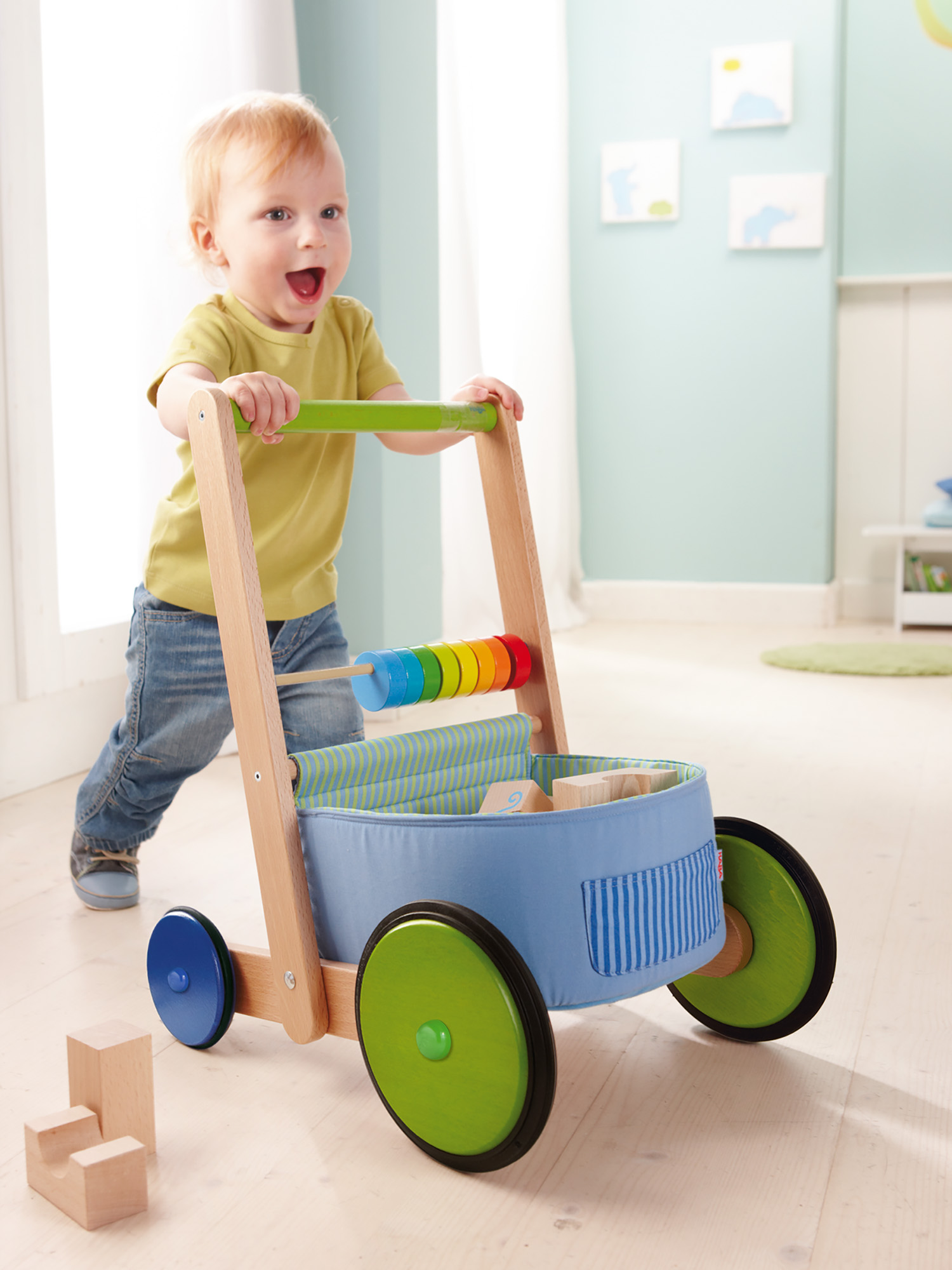 Un bambino piccolo che spinge un girello di legno con giocattoli. Il girello ha perline colorate e tessuto blu. Il bambino sorride.