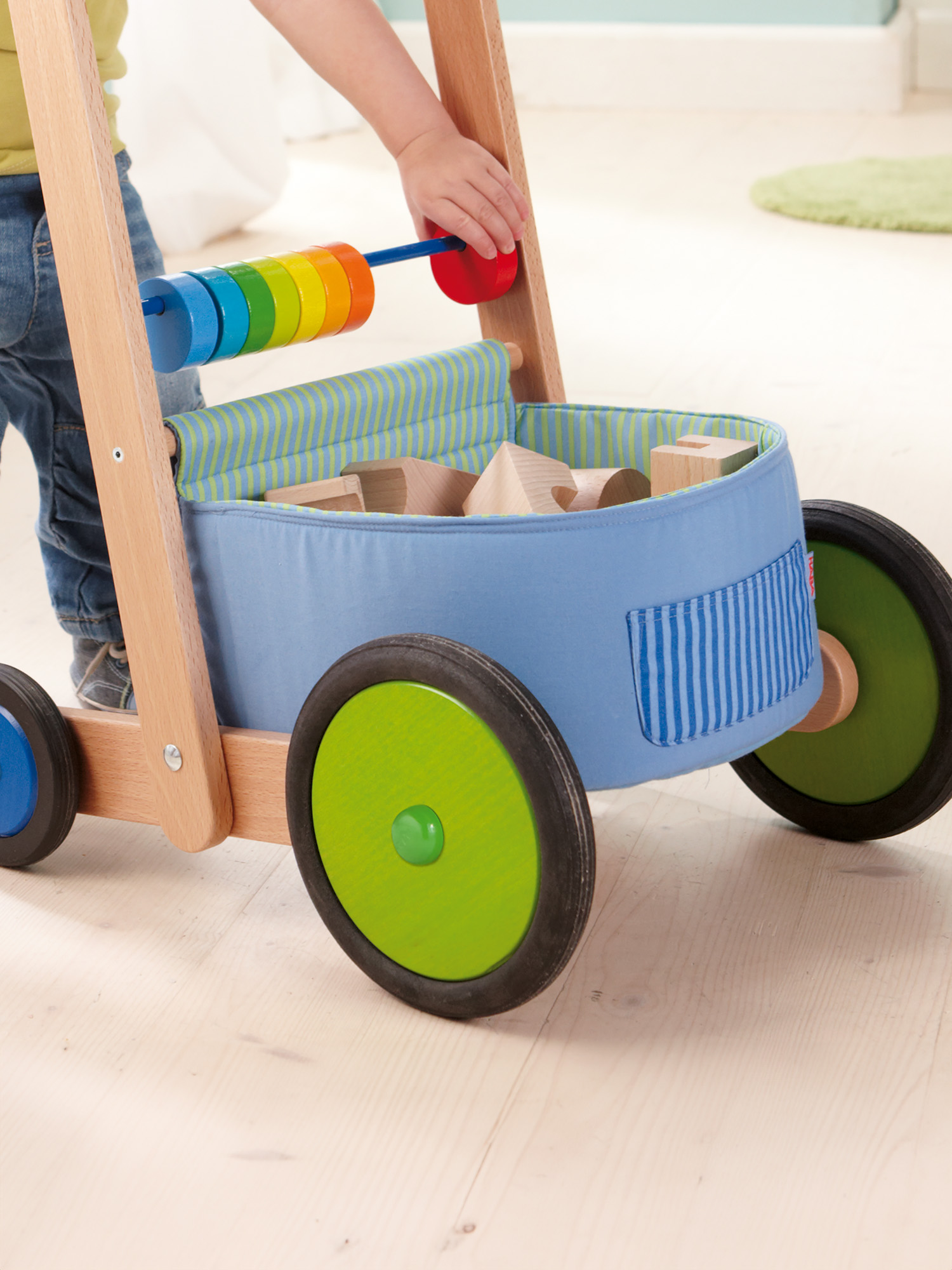 Un bambino gioca con un girello di legno. Il girello ha un contenitore blu pieno di blocchi e anelli colorati.