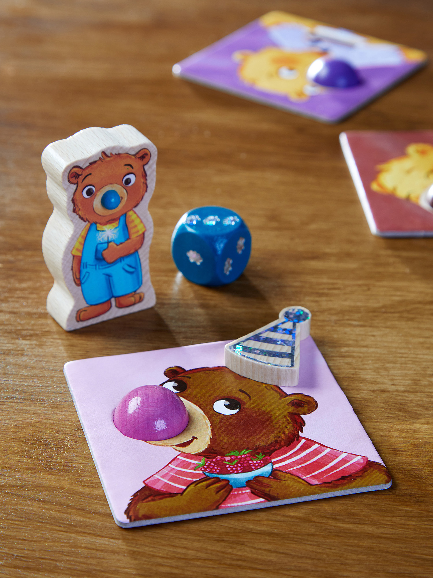 Una figura di orso in legno, un dado blu e carte a tema orso su una superficie di legno. L'orso indossa una tuta e ha un ciuccio.