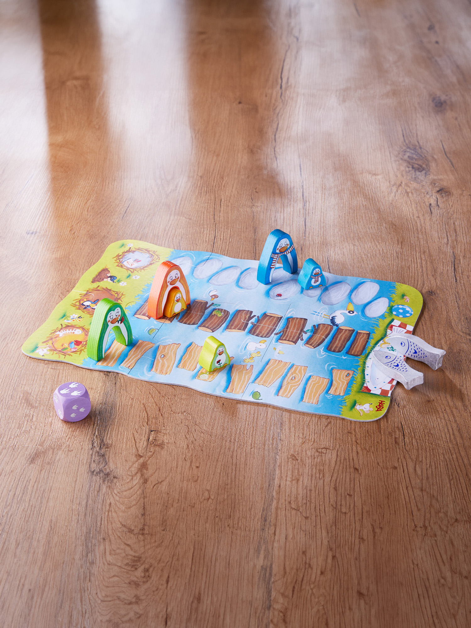 Ein Brettspiel mit pinguinförmigen Figuren auf einer Holzoberfläche. Ein Würfel ist vorhanden, und das Spielbrett zeigt eine Flussszene.