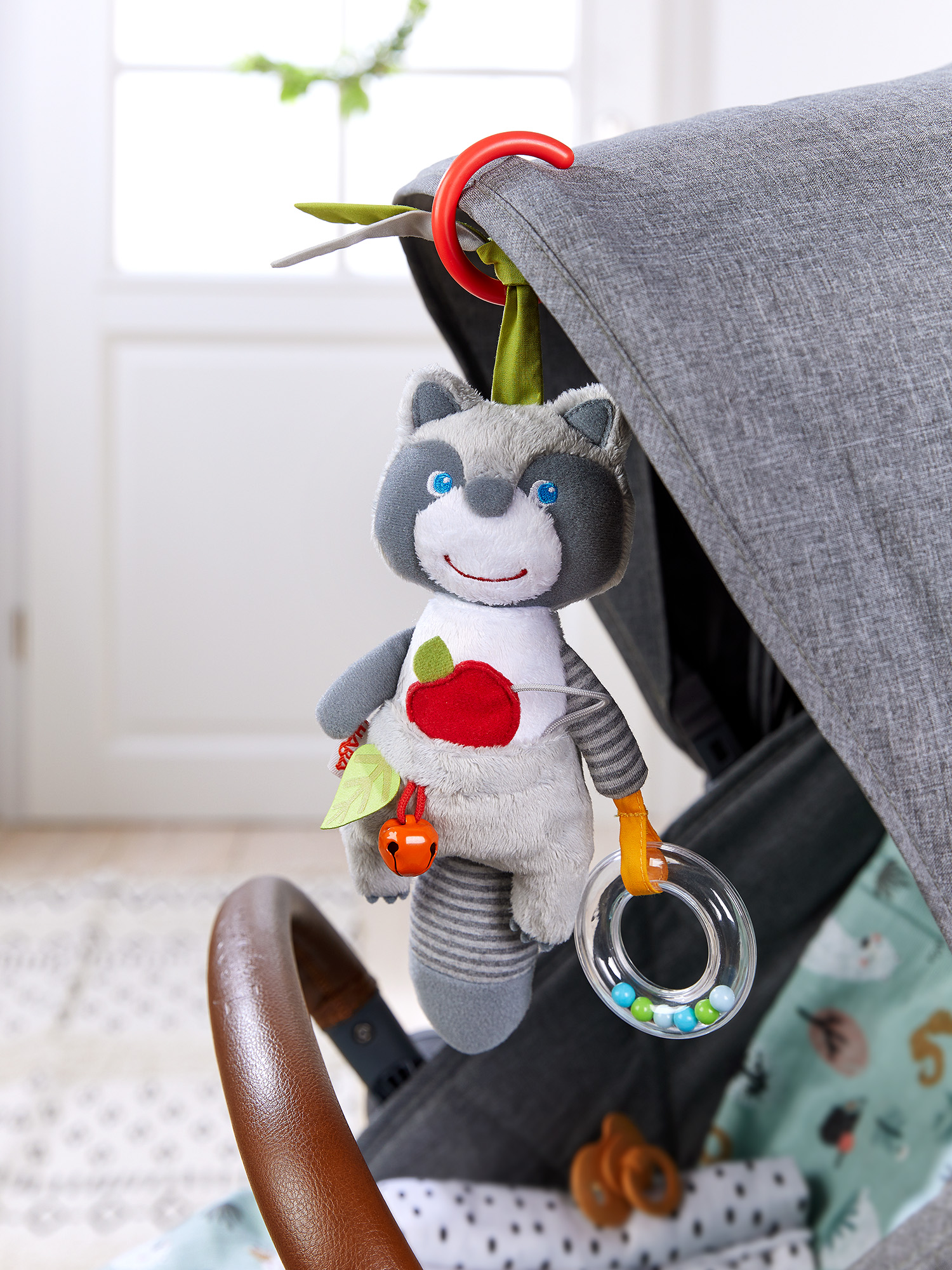 Ein graues Kuscheltier-Waschbär hängt an einem Kinderwagen. Es hat einen roten Apfel, eine Glocke und eine durchsichtige Rassel. Der Kinderwagen ist grau.